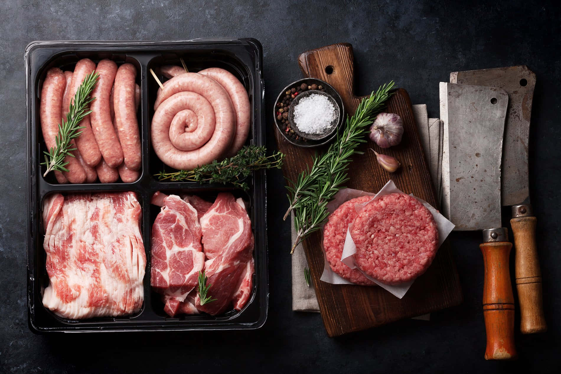 meat 桌布 壁紙 — 從上方俯瞰的視角，展示生肉，包括香腸、肉餅和培根，與香草、鹽、大蒜和剁刀一同排列在深色紋理表面上。