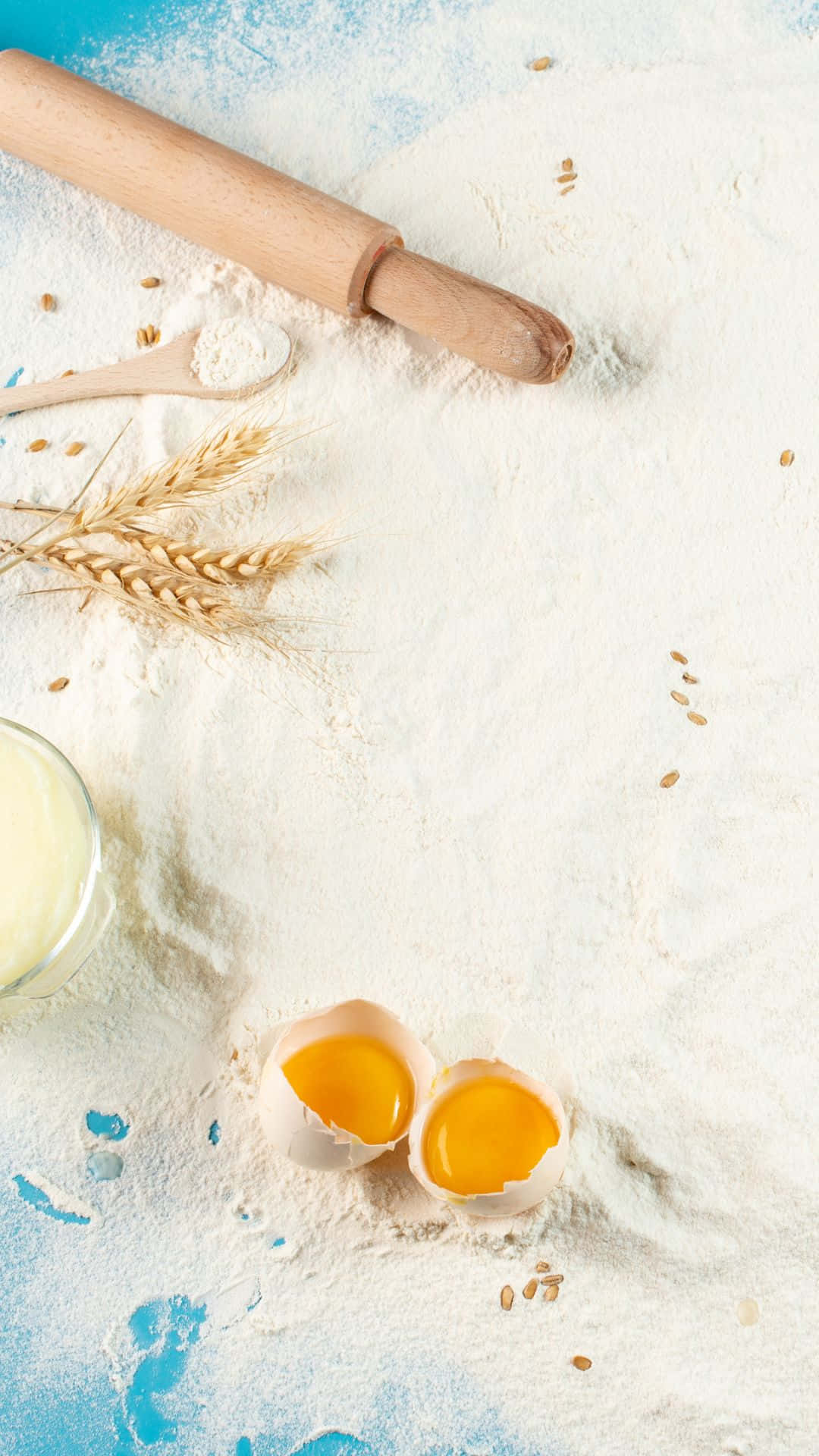 Egg 桌布 壁紙 — 從上方俯瞰的視角，裂開的雞蛋蛋黃呈現亮黃色，靜置在麵粉中，旁邊有擀麵棍、小麥莖和牛奶罐，置於帶紋理的藍色表面上。