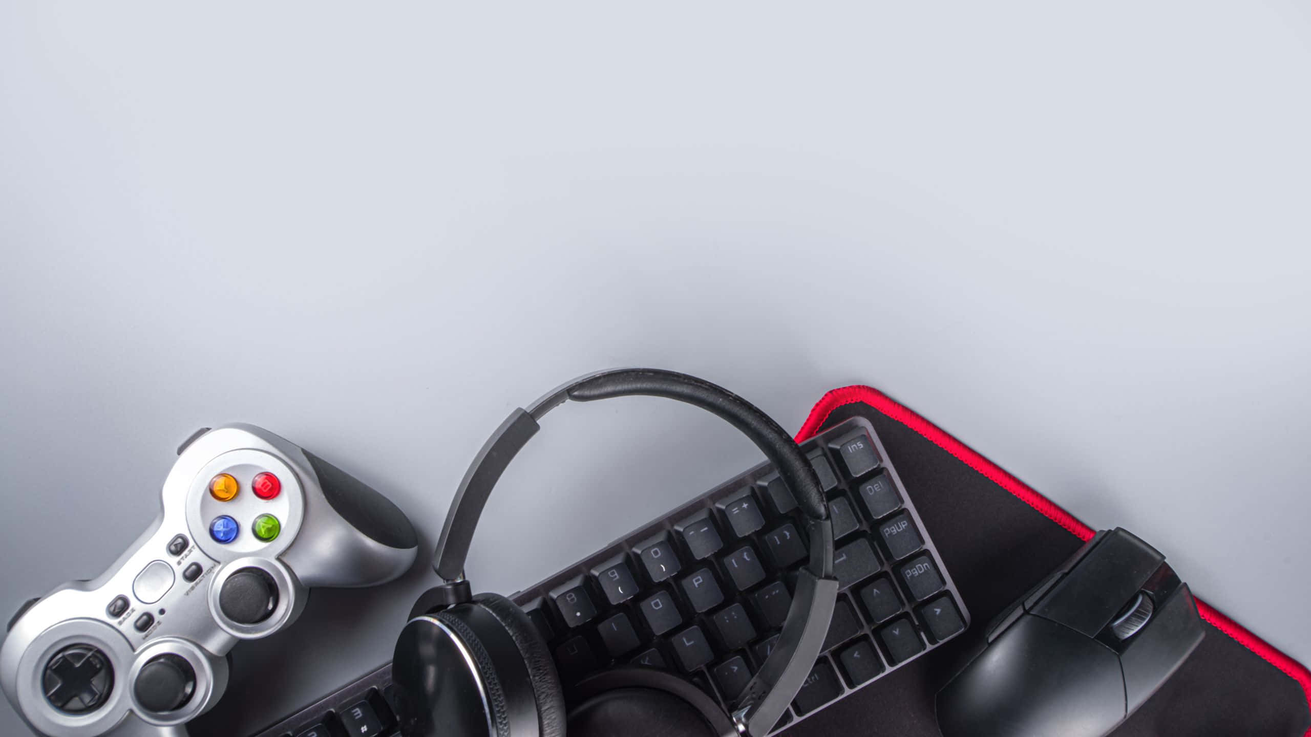 Gaming Banner 桌布 壁紙 — 從上方俯瞰遊戲裝備，包括控制器、耳機、鍵盤和滑鼠，放在紅色邊緣的滑鼠墊上，背景為淺灰色。