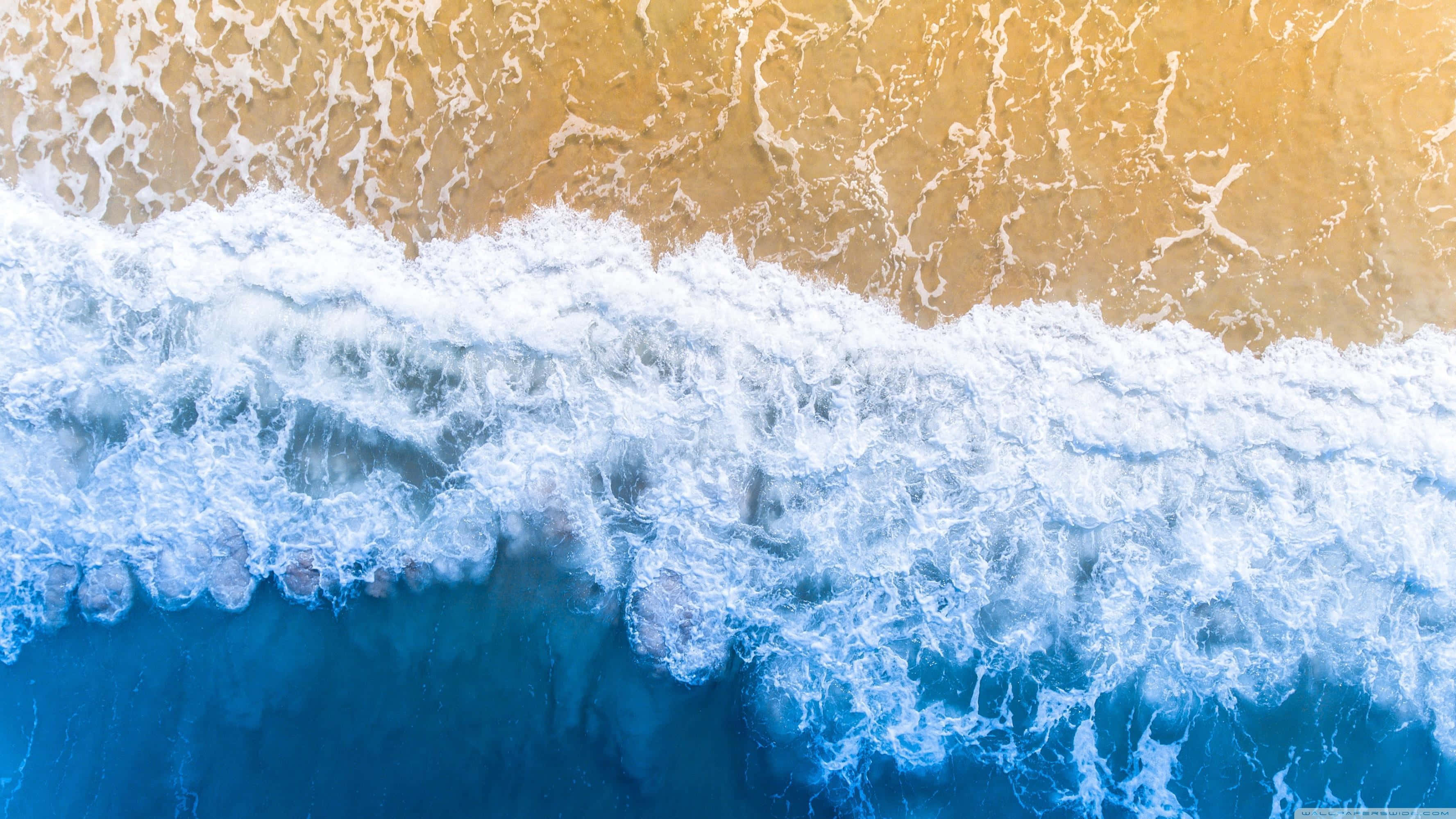 waves 桌布 壁紙 — 從上空俯瞰海浪拍打金色沙灘，泛起白色泡沫的景色。