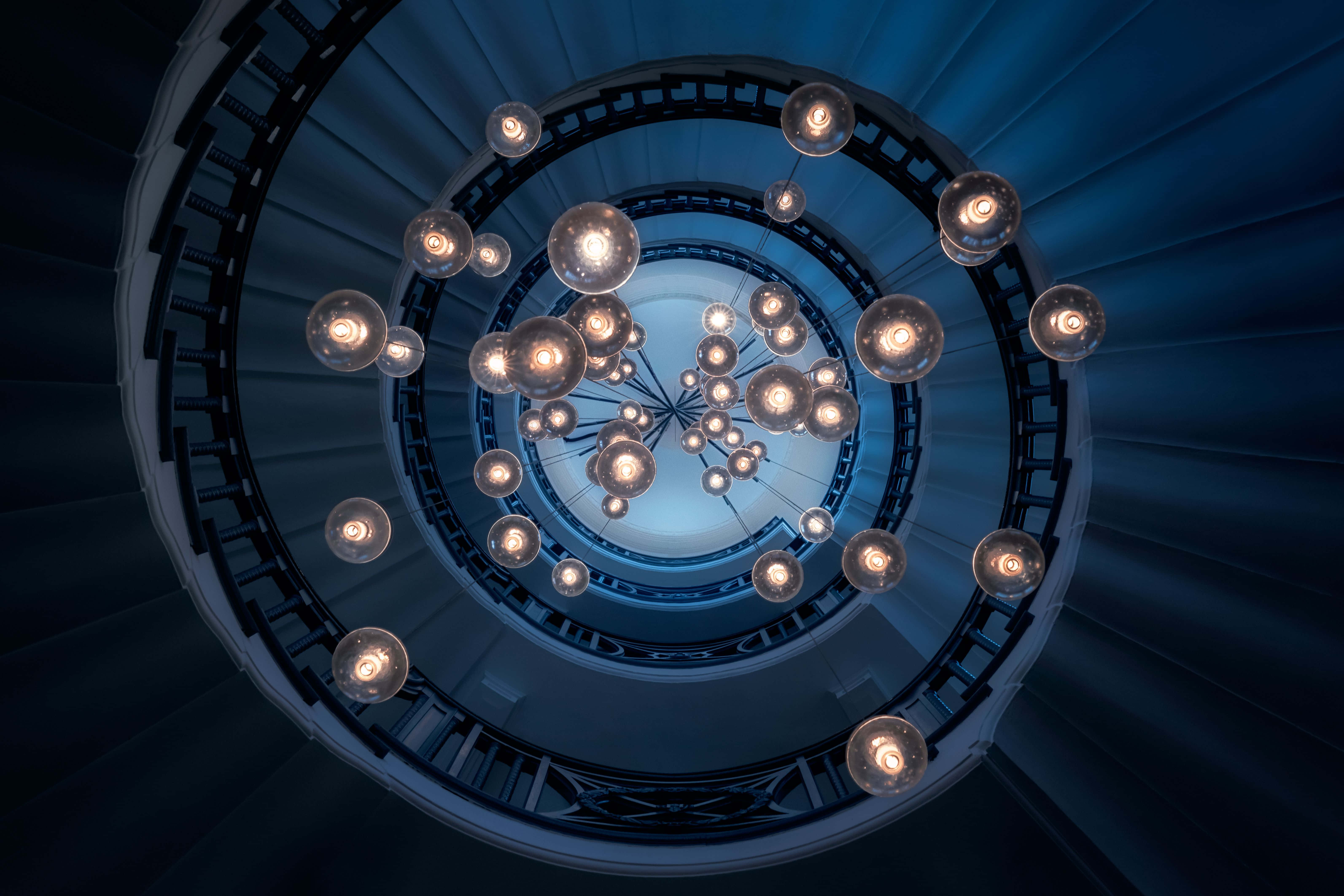 stairs 桌布 壁紙 — 從下方仰望的壯觀螺旋樓梯，懸掛著發光的球形燈飾，處於現代建築環境中。
