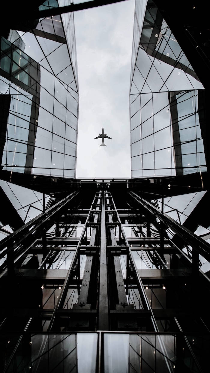 Skyscraper 桌布 壁紙 — 從低角度仰望現代玻璃摩天大樓的反光外牆，看到一架遙遠的飛機在頭頂飛過。