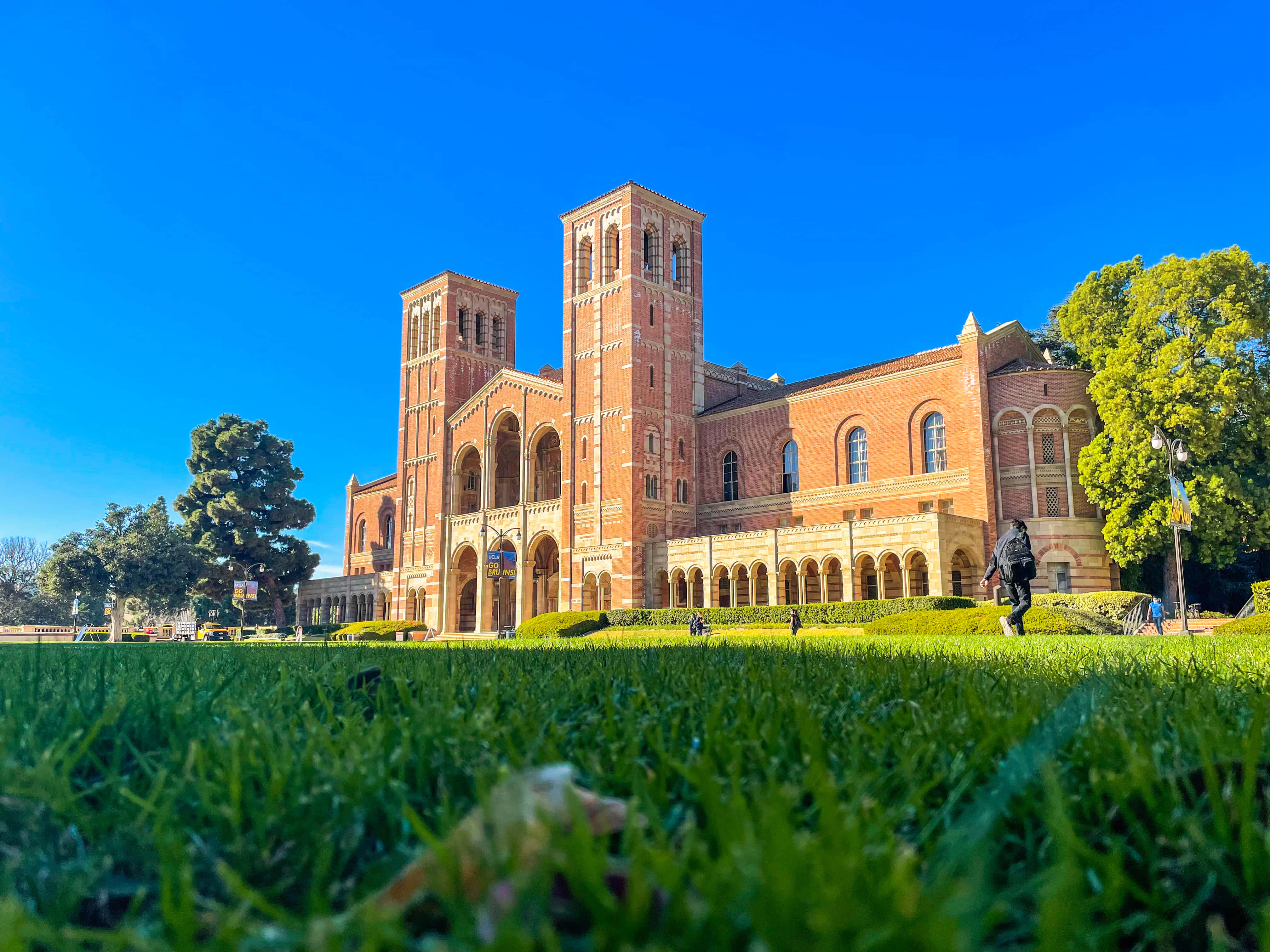 Ucla Campus 桌布 壁紙 — 從低角度觀看一座宏偉的紅磚大學建築，擁有雙塔，在清澈的藍天下，被充滿活力的綠色草地和樹木環繞。