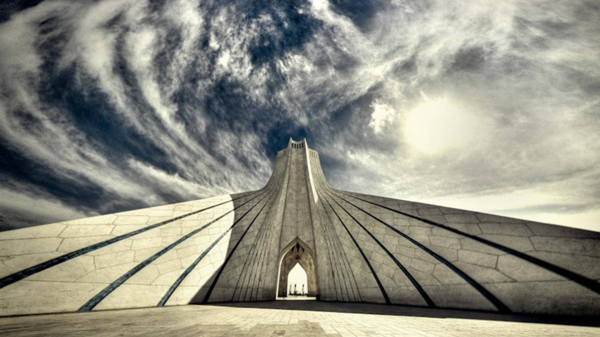 Tehran 桌布 壁紙 — 從低角度觀看德黑蘭的阿扎迪塔，頭頂有戲劇性的漩渦雲和明亮的太陽。