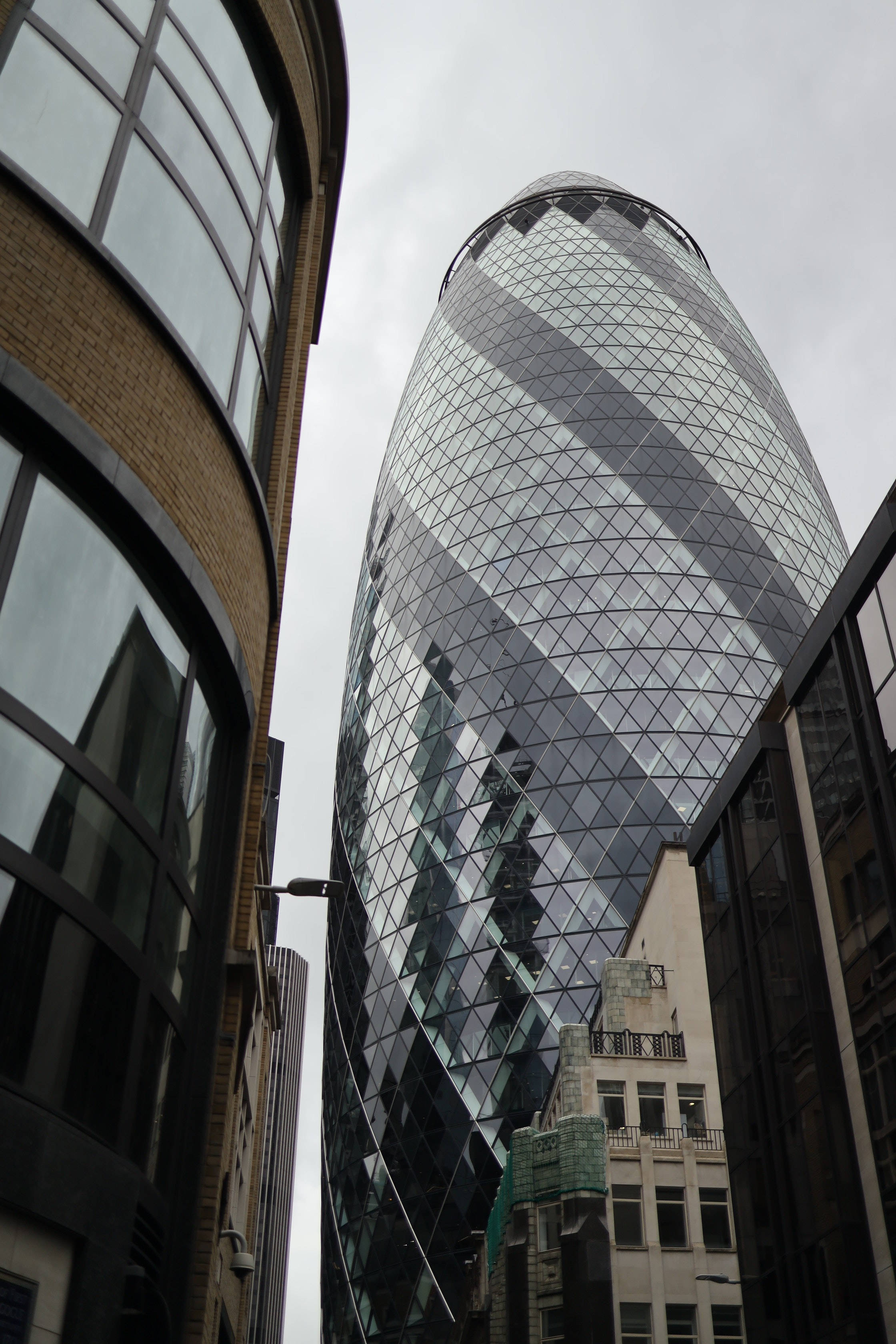 Buildings 桌布 壁紙 — 從低角度觀看標誌性的Gherkin摩天大樓，其玻璃外牆反射灰色雲層，周圍環繞著城市建築。