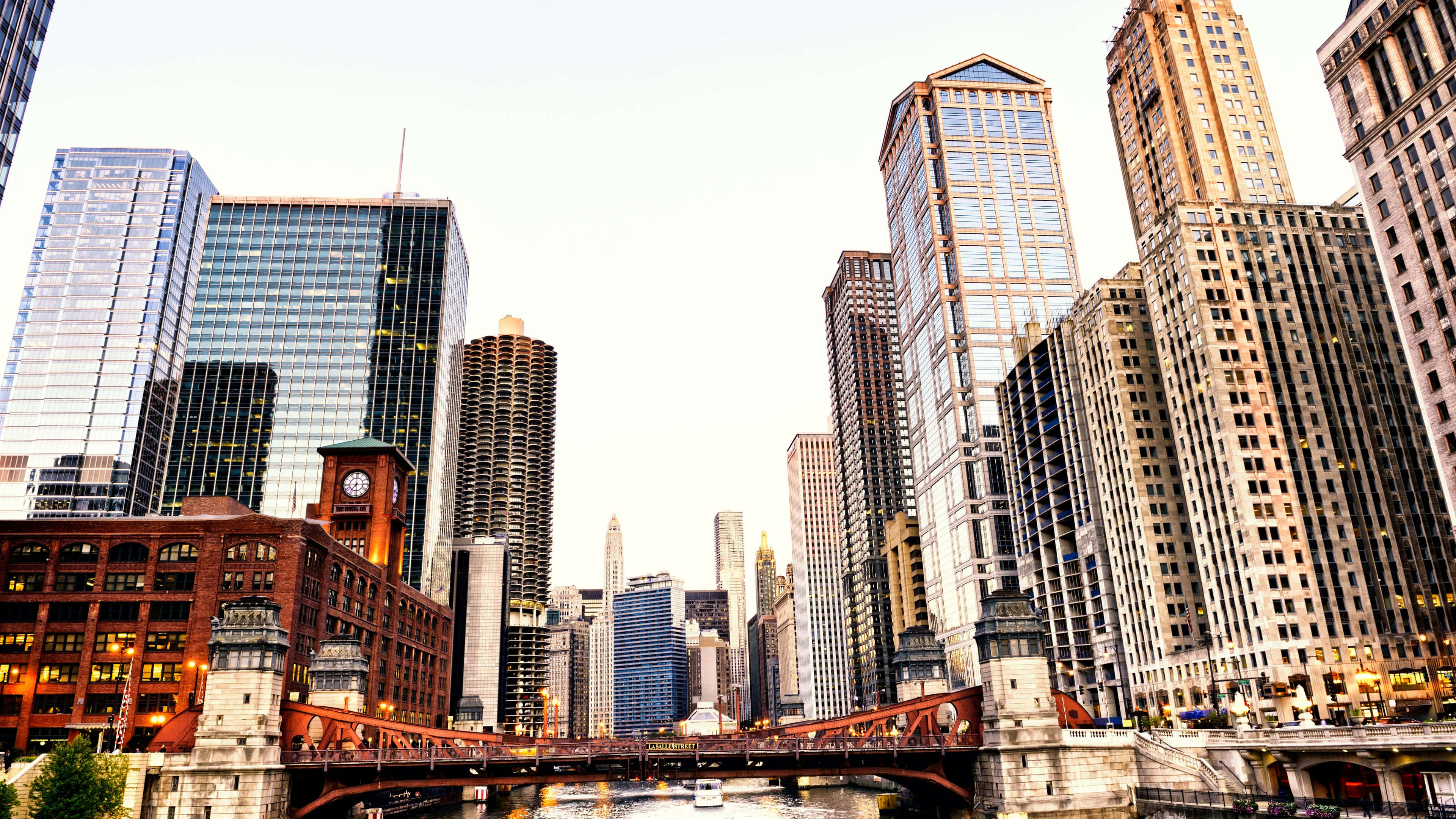 4K Ultra HD Chicago 桌布 壁紙 — 從低角度觀賞芝加哥黃昏時分的天際線，一條紅色橋樑橫跨河流，兩旁是高聳的玻璃和磚造摩天大樓。