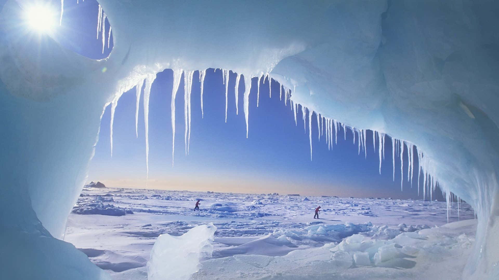 North Pole 桌布 壁紙 — 從冰洞內部欣賞的壯麗景色，懸掛的冰柱框住了明亮的藍天和兩個穿越冰凍廣闊區域的人影。