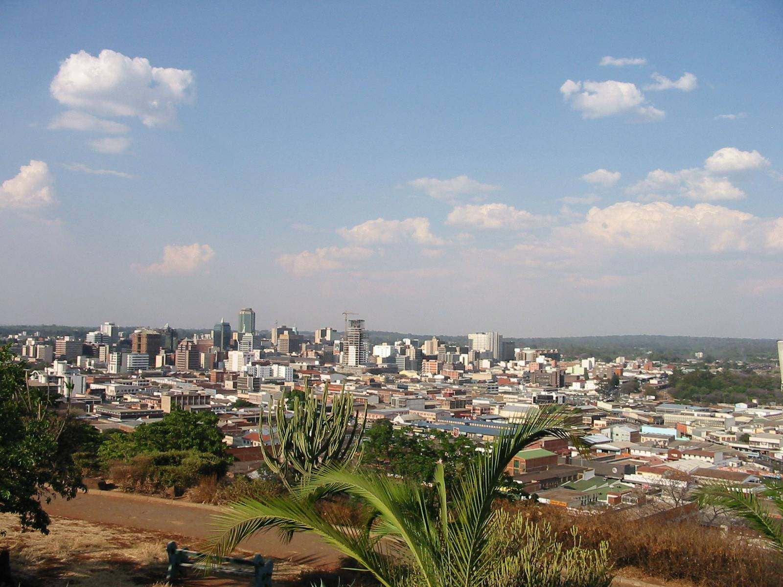 Zimbabwe 桌布 壁紙 — 從前景有熱帶植物的山坡上，眺望廣闊的城市天際線，上方是明亮的藍天，點綴著蓬鬆的白雲。