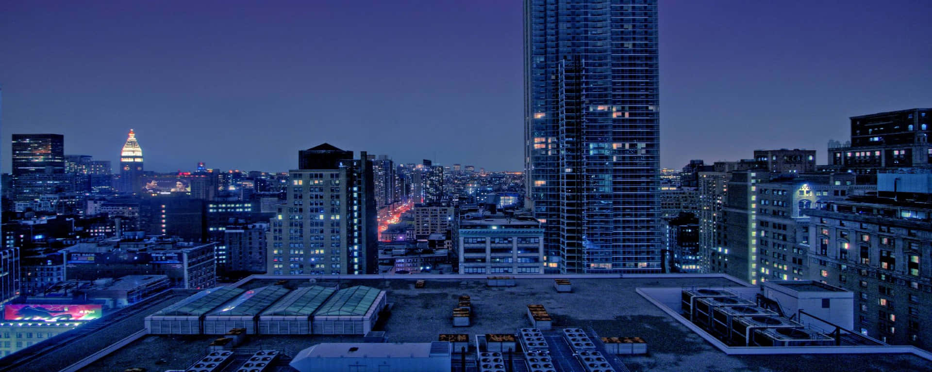 城市風景 桌布 壁紙 — 從屋頂俯瞰黃昏時分的城市天際線，窗戶閃爍著光芒，遠處的塔樓在深紫色天空下被照亮。