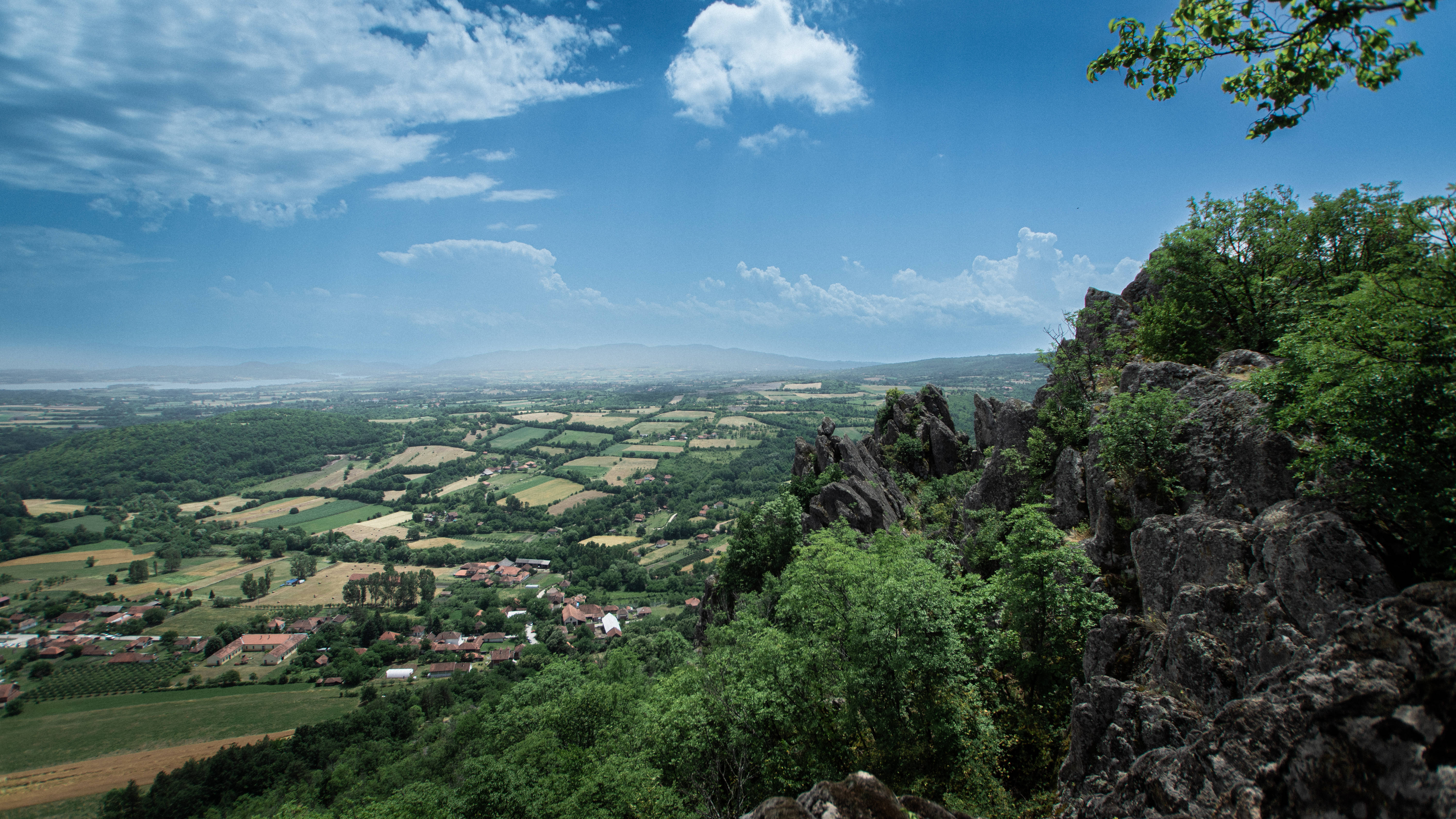 Serbia 桌布 壁紙 — 從岩石懸崖俯瞰的 panoramic 景觀，翠綠的山谷中有拼布般的田野和散落的村莊，明亮的藍天下有蓬鬆的雲朵。