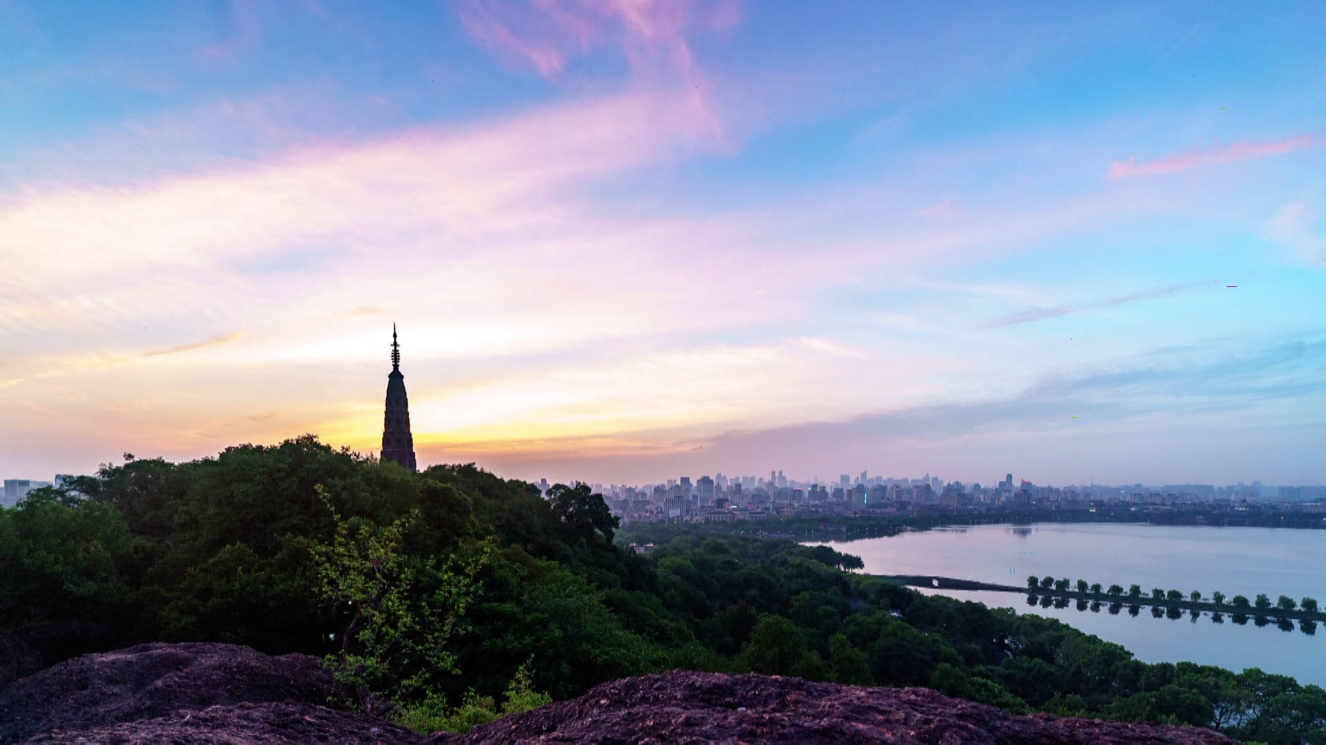 Hangzhou 桌布 壁紙 — 從岩石觀景點欣賞寧靜的日落景色，顯示一座歷史悠久的寶塔坐落在鬱鬱蔥蔥的山丘上，俯瞰城市天際線和寧靜的湖泊。