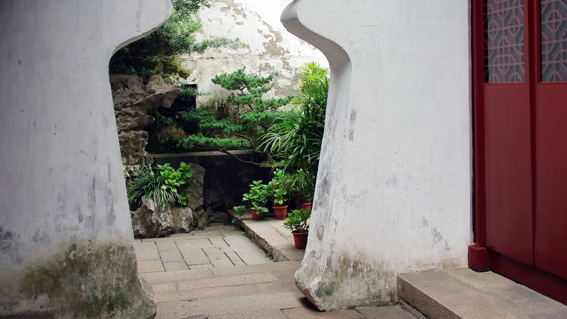Suzhou 桌布 壁紙 — 從彎曲的白色石拱門望出去，一個傳統的中國花園，遠處有盆栽植物和長滿苔蘚的石頭。