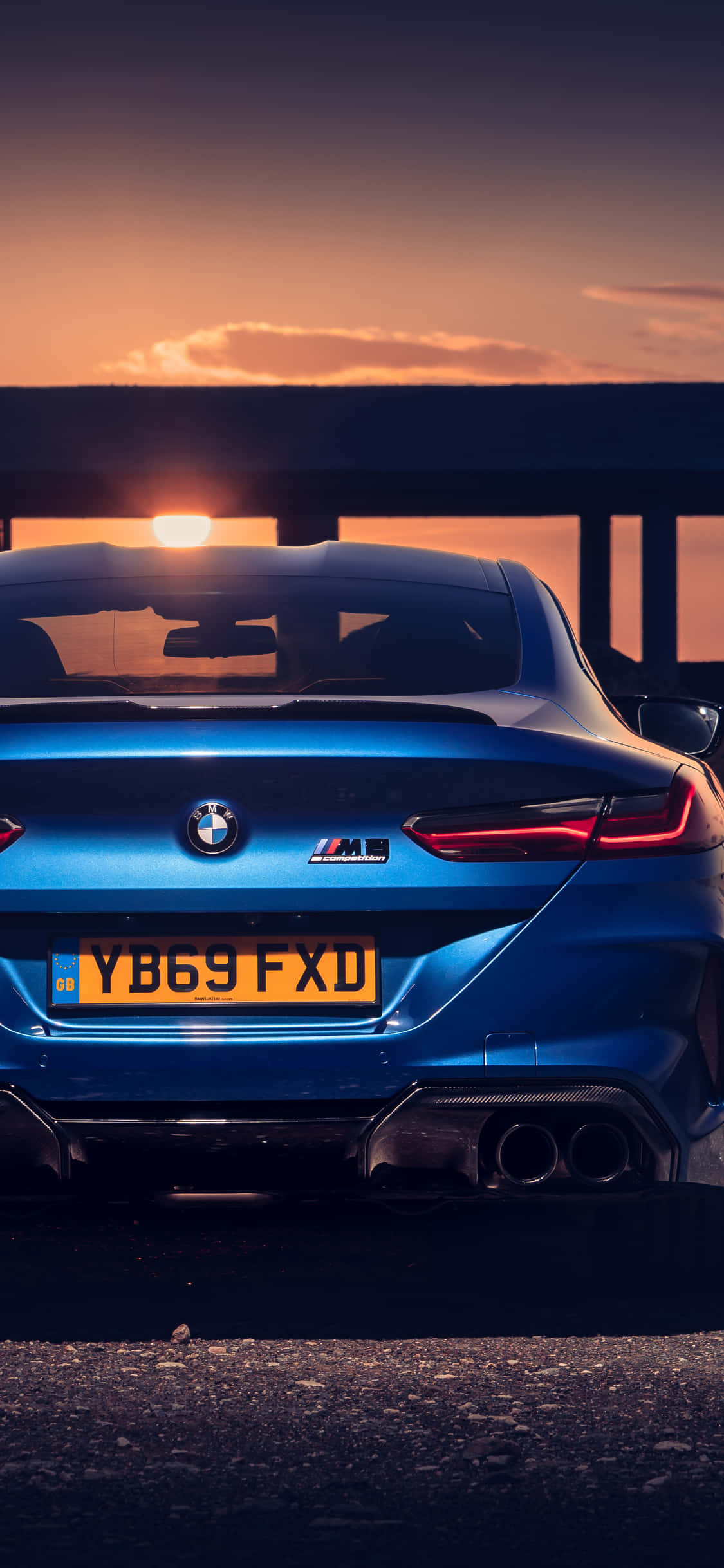 Bmw iPhone 桌布 壁紙 — 從後方觀看，一輛時尚的藍色BMW M8 Coupe在日落時分，帶有發光的尾燈和溫暖的天空。