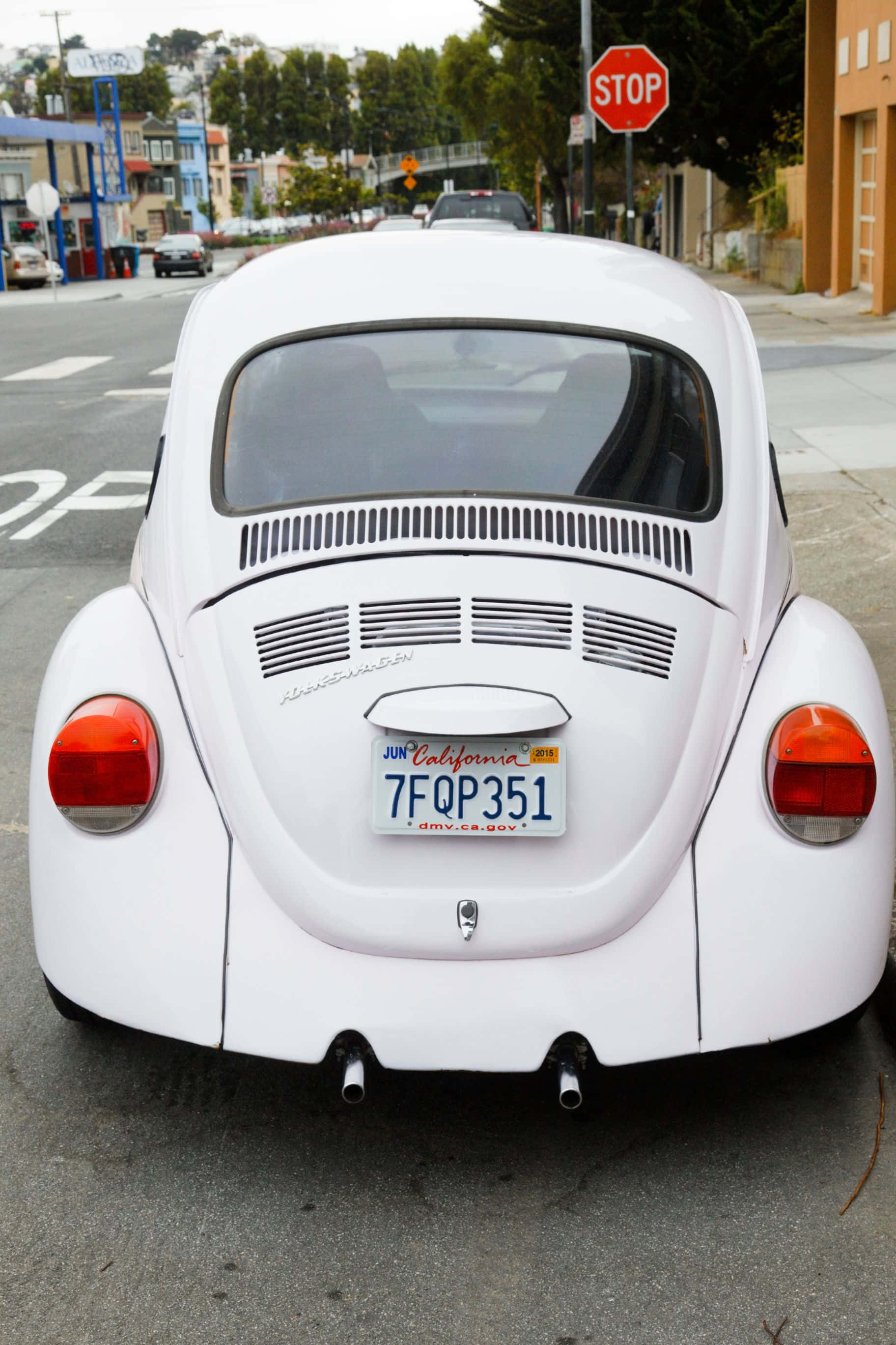 1824x2736 桌布 壁紙 — 從後方觀看一輛完美無瑕的白色 vintage Volkswagen Beetle 停泊在城市的街道上，背景有停車標誌和色彩繽紛的建築物。
