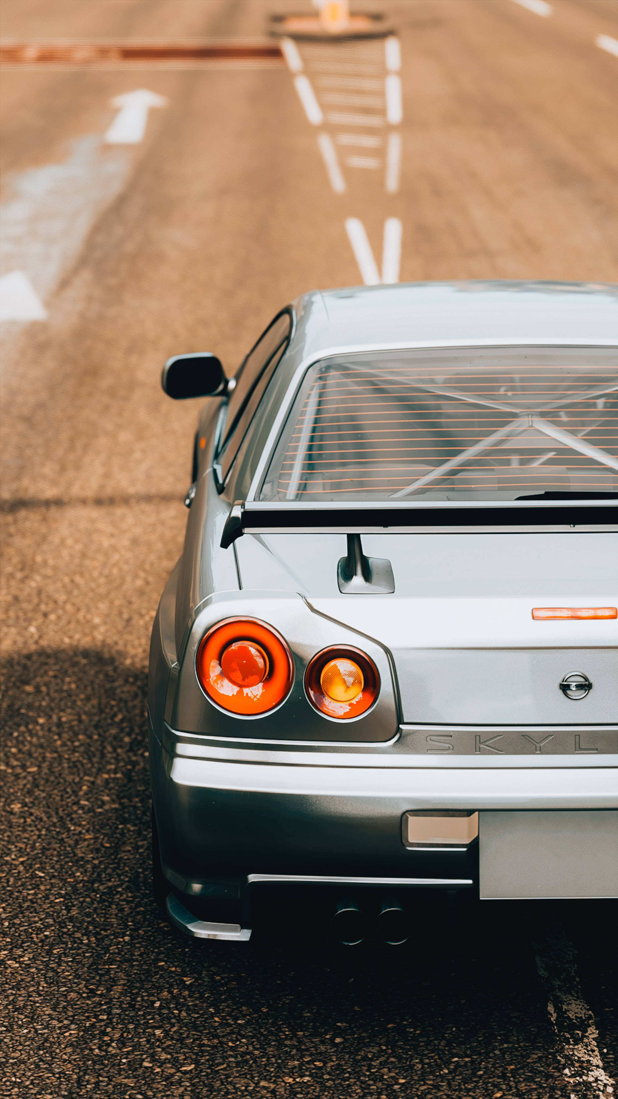 Nissan 桌布 壁紙 — 從後方觀看一輛銀色日產Skyline R32跑車，行駛在陽光普照的瀝青路上，帶有白色車道標線。