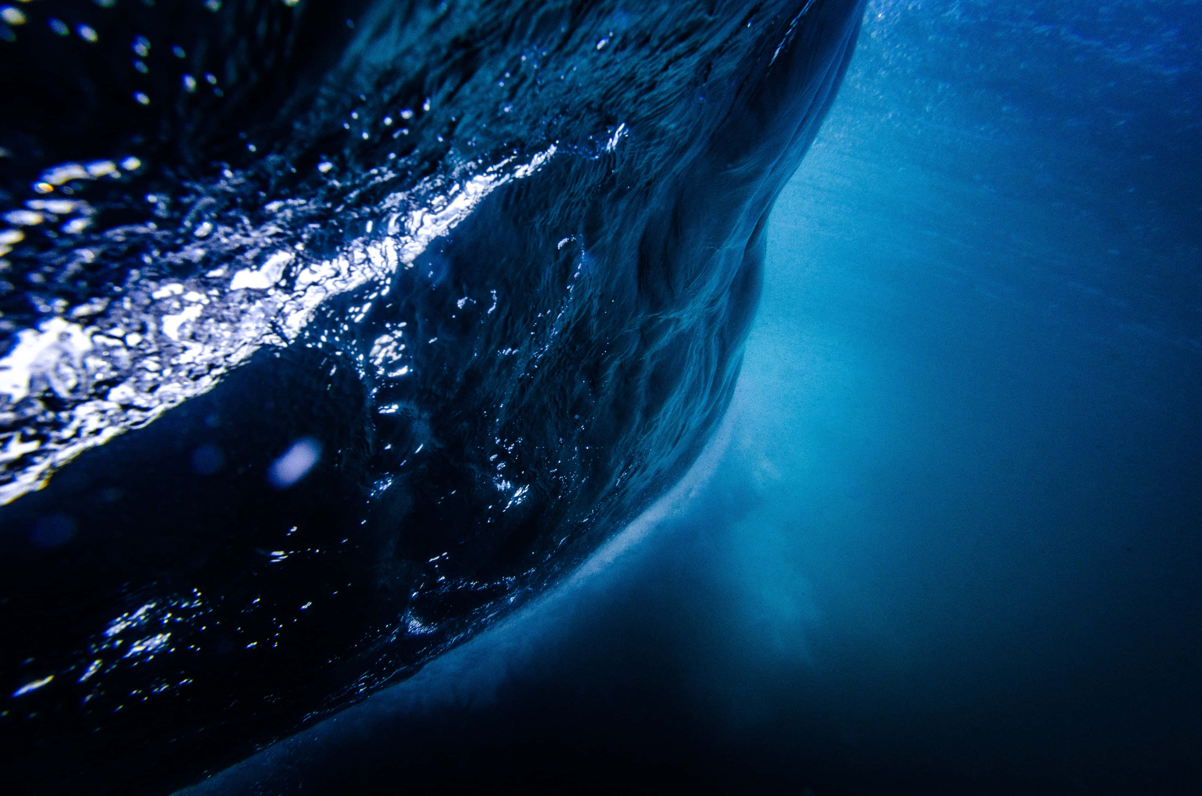 Underwater Ocean 桌布 壁紙 — 從捲曲的海浪下方拍攝的戲劇性水下景觀，陽光照射的藍色海水和閃閃發光的表面紋理。