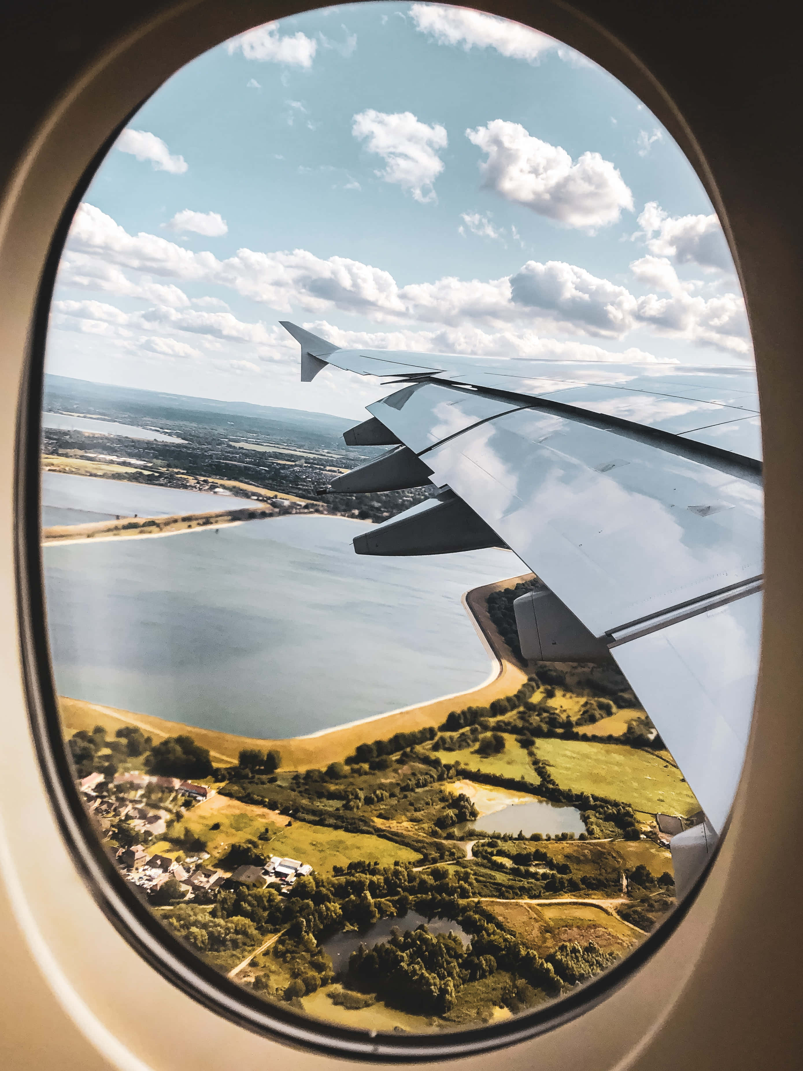 Airplane Window 桌布 壁紙 — 從橢圓形飛機窗戶看到的飛機機翼，俯瞰綠色田野、湖泊和散落的城鎮，天空明亮多雲。