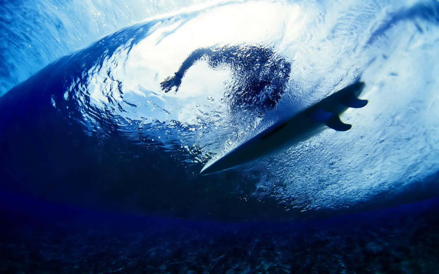 surfing 桌布 壁紙 — 從水下視角，陽光穿透捲曲的藍色海水，一名衝浪者在巨大的海浪中衝浪。