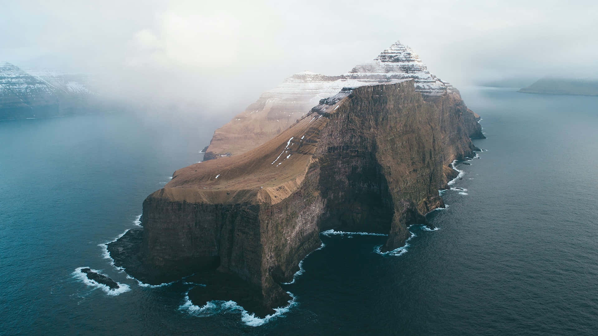 Awe-inspiring 桌布 壁紙 — 從深藍色海洋中升起的雪覆蓋懸崖島嶼的鳥瞰圖，薄霧在其崎嶇邊緣盤旋。