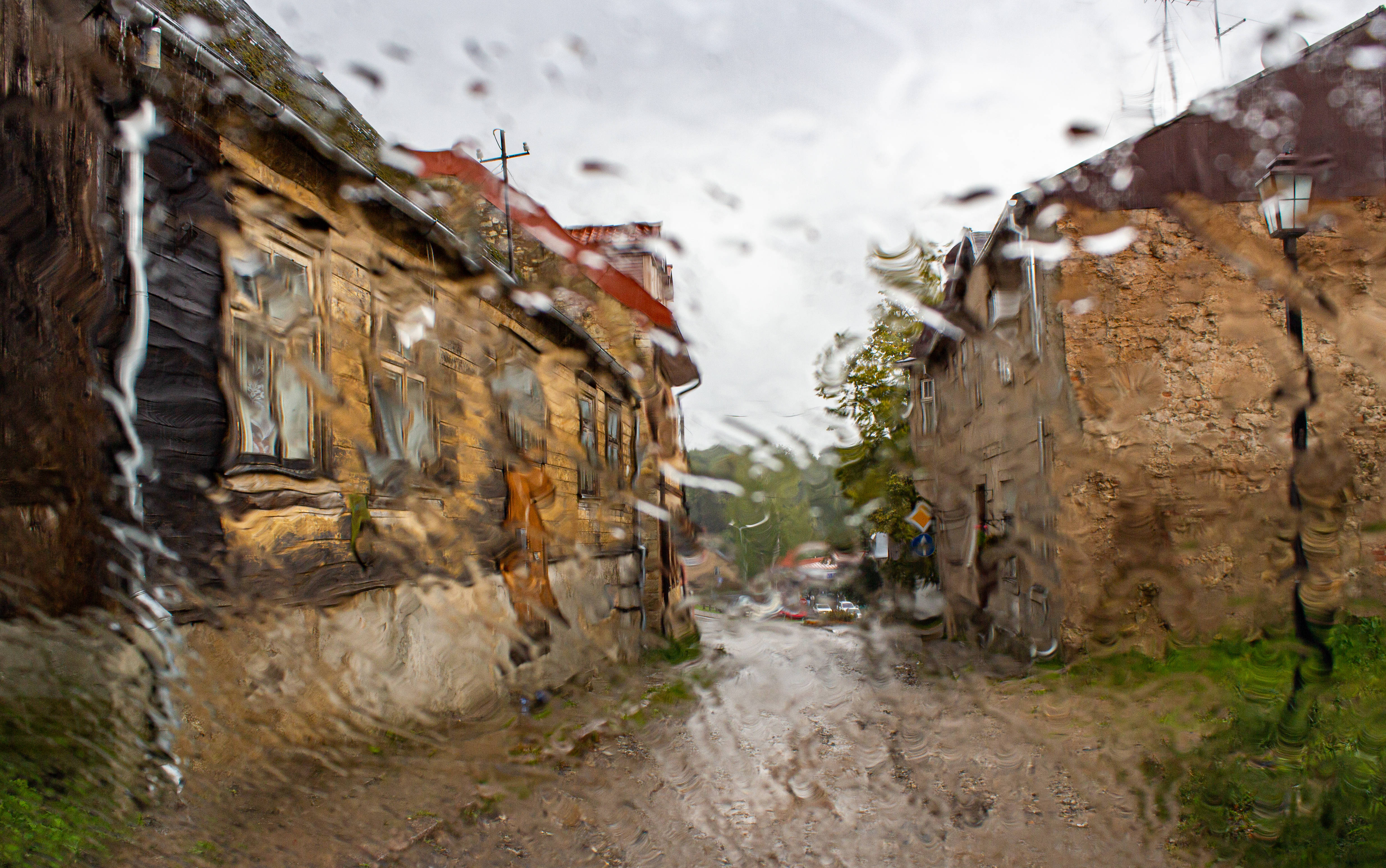 Latvia 桌布 壁紙 — 從濺滿雨點的窗戶望出去，一條狹窄泥濘的村莊街道，兩旁是風化的石頭和木頭房屋，天空陰沉。