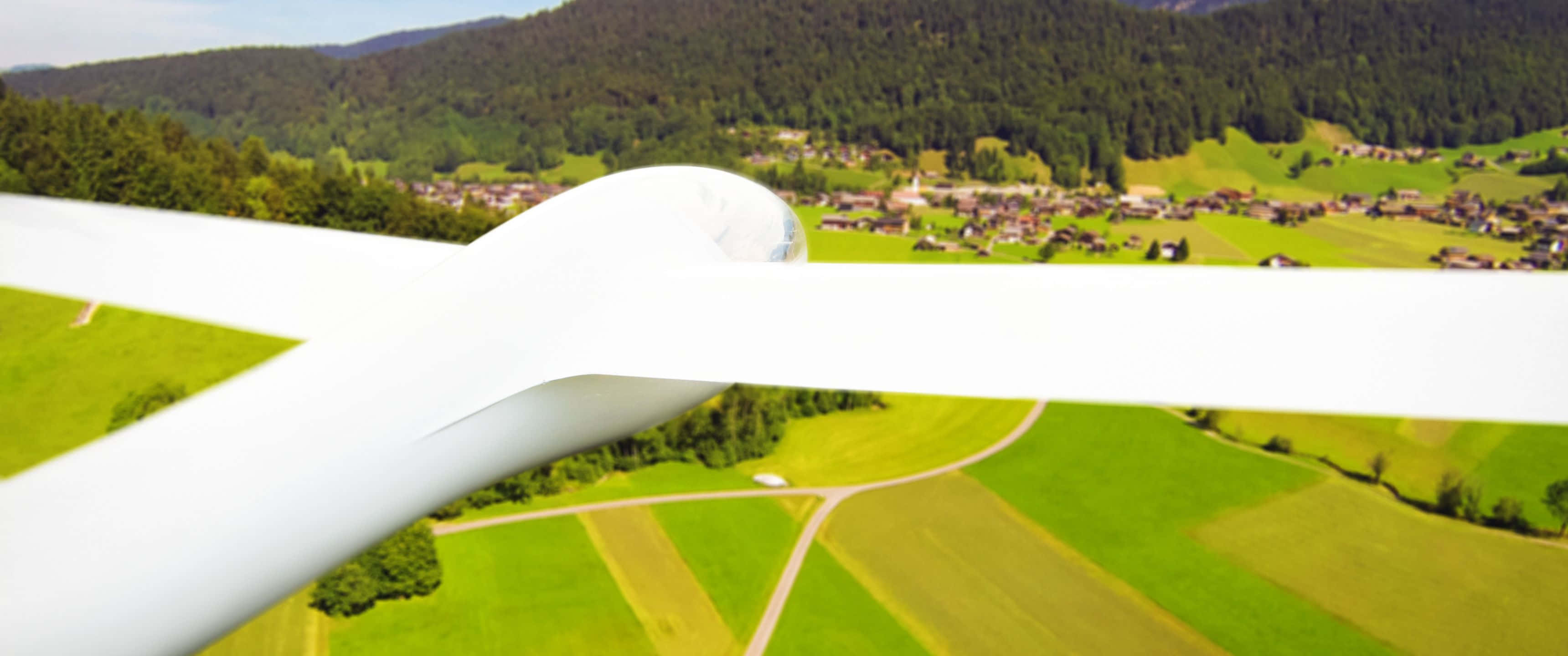 3440x1440p Small Planes 桌布 壁紙 — 從白色滑翔機俯瞰，翱翔於充滿活力的綠色田野和森林覆蓋的山谷，下方有一個村莊。