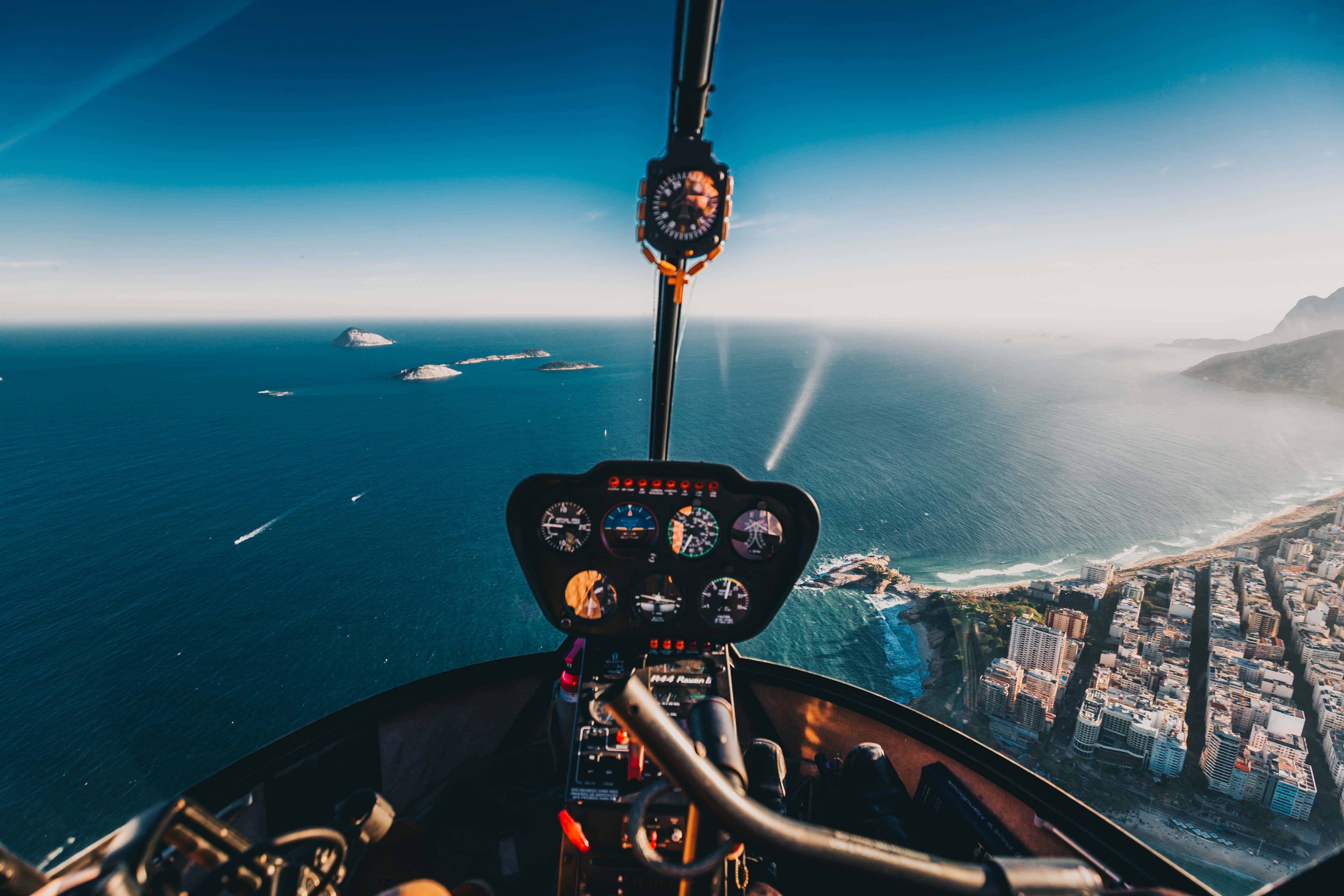 Helicopter 桌布 壁紙 — 從直升機駕駛艙以第一人稱視角俯瞰沿海城市，碧綠海洋與霧氣繚繞的山脈在清澈藍天下展開。