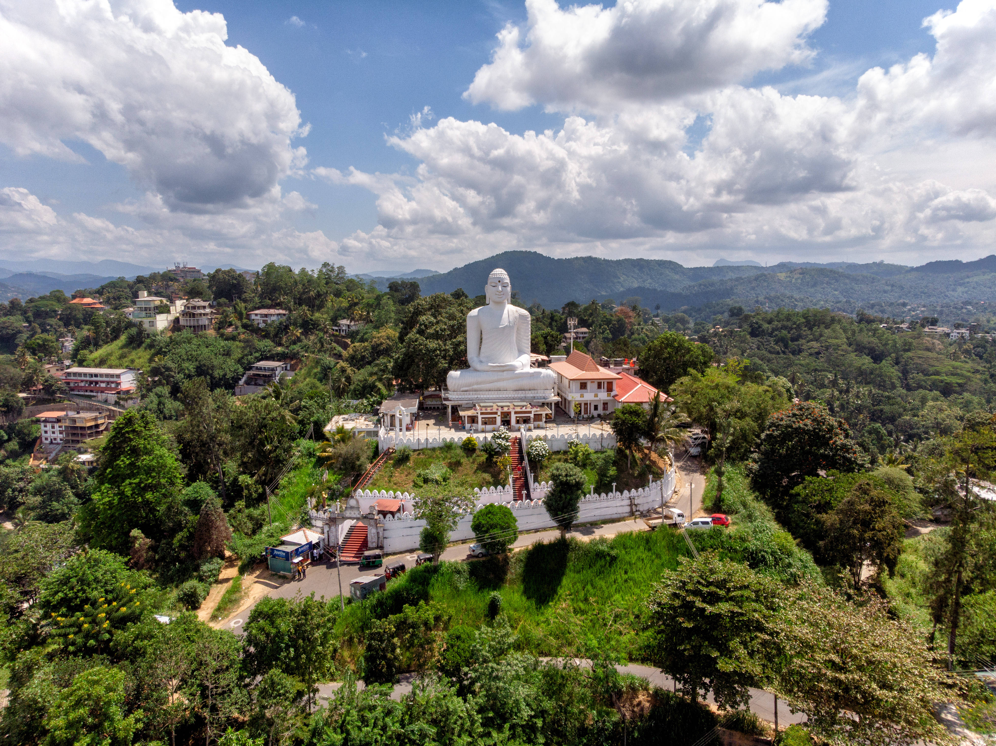Gautam Buddha 桌布 壁紙 — 從空中俯瞰，一座巨大的白色佛像矗立在鬱鬱蔥蔥的山頂上，俯瞰著城鎮和遠處的山脈，天空部分多雲。