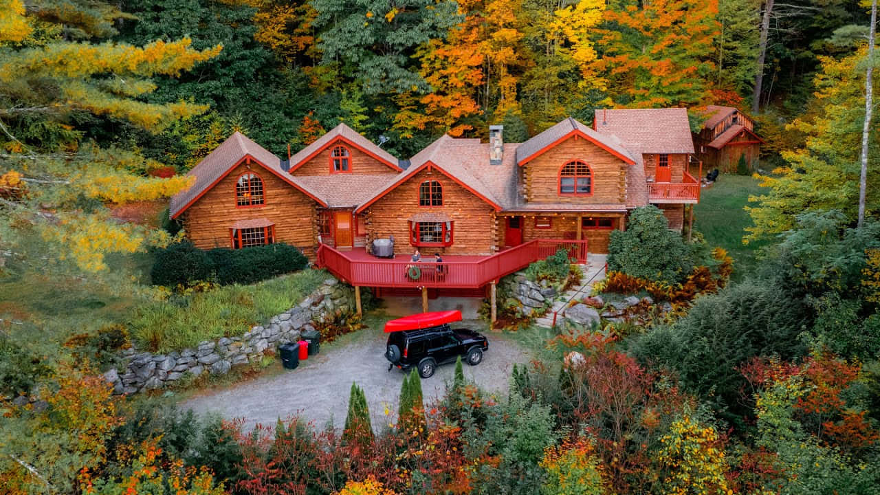 Fall Cabin 桌布 壁紙 — 從空中俯瞰，一座帶有紅色飾邊的質樸木屋坐落在色彩繽紛的秋林中。