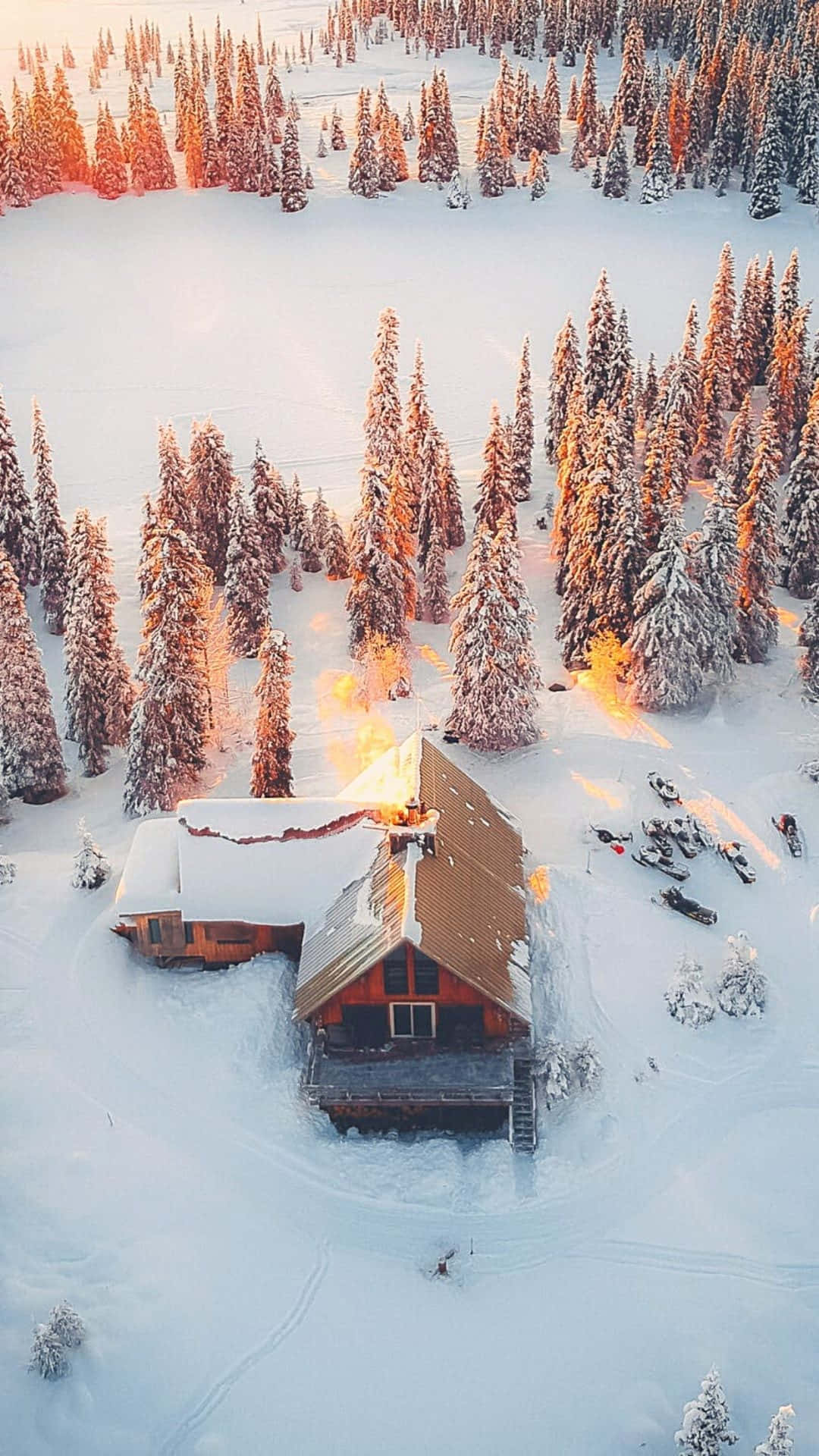 Cute Winter Phone 桌布 壁紙 — 從空中俯瞰，一間舒適的木屋坐落在白雪覆蓋的森林中，沐浴在金色的夕陽餘暉裡。
