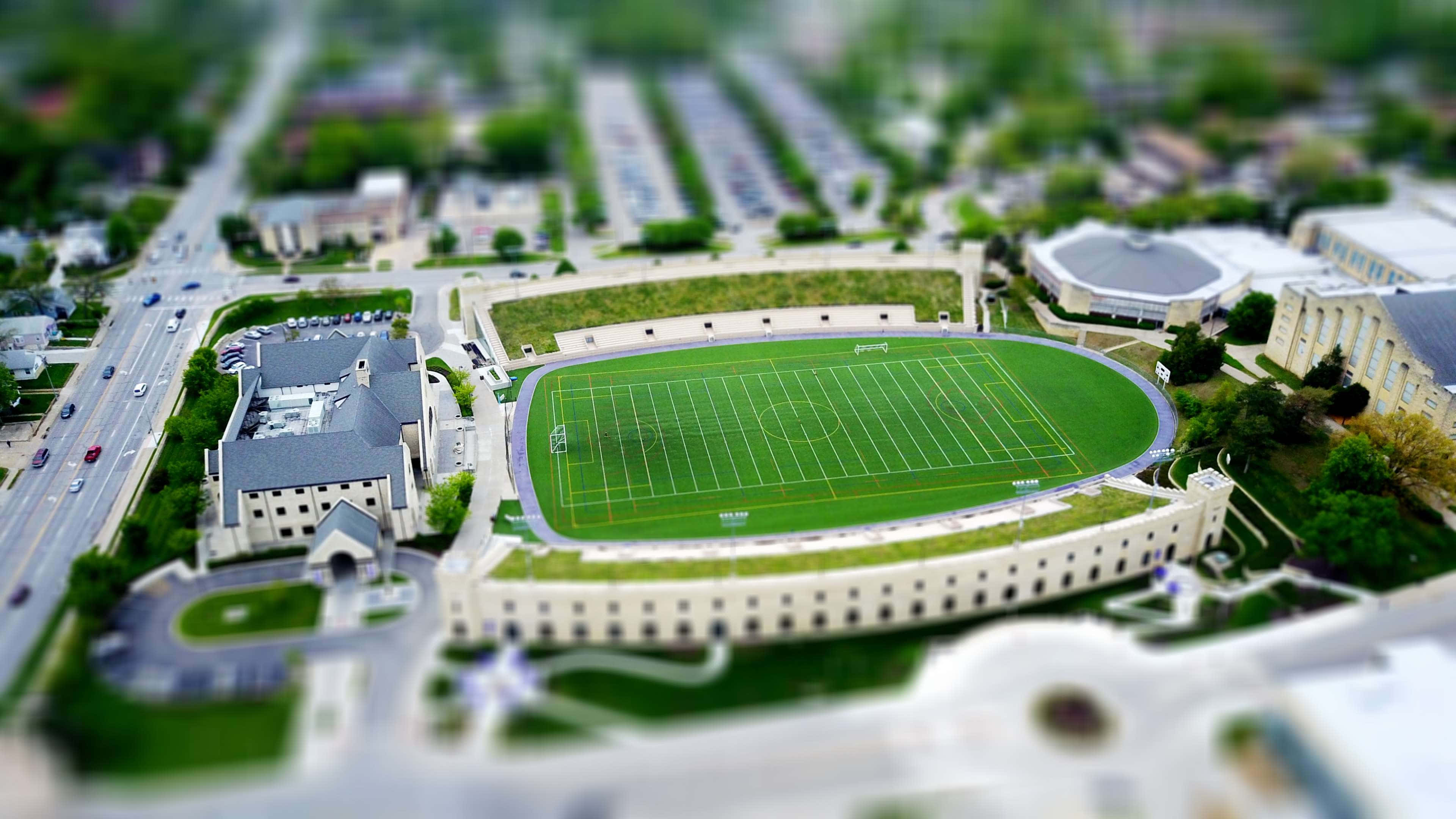 Football Field 桌布 壁紙 — 從空中俯瞰，以移軸效果呈現的翠綠足球場，四周環繞著校園建築和道路。
