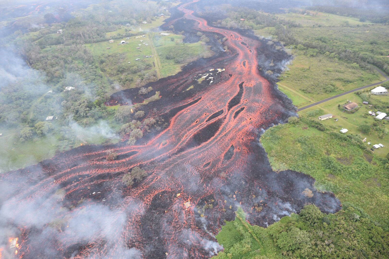 Volcano 桌布 壁紙 — 從空中俯瞰，熾熱的紅色熔岩流穿過鬱鬱蔥蔥的綠色景觀，煙霧在散落的房屋附近升起。