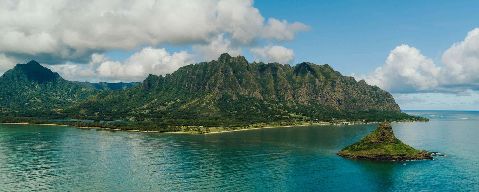 Hawaiian Island 桌布 壁紙 — 從空中俯瞰，鬱鬱蔥蔥的綠色山脈從碧綠的海水中升起，前景有一個小島，天空部分多雲。