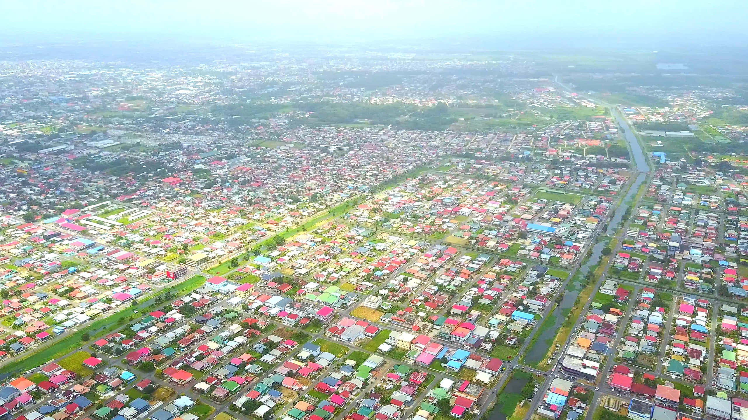 Suriname 桌布 壁紙 — 從空中俯瞰一個廣闊的郊區社區，色彩繽紛的屋頂和蜿蜒的道路在柔和的晨光下。