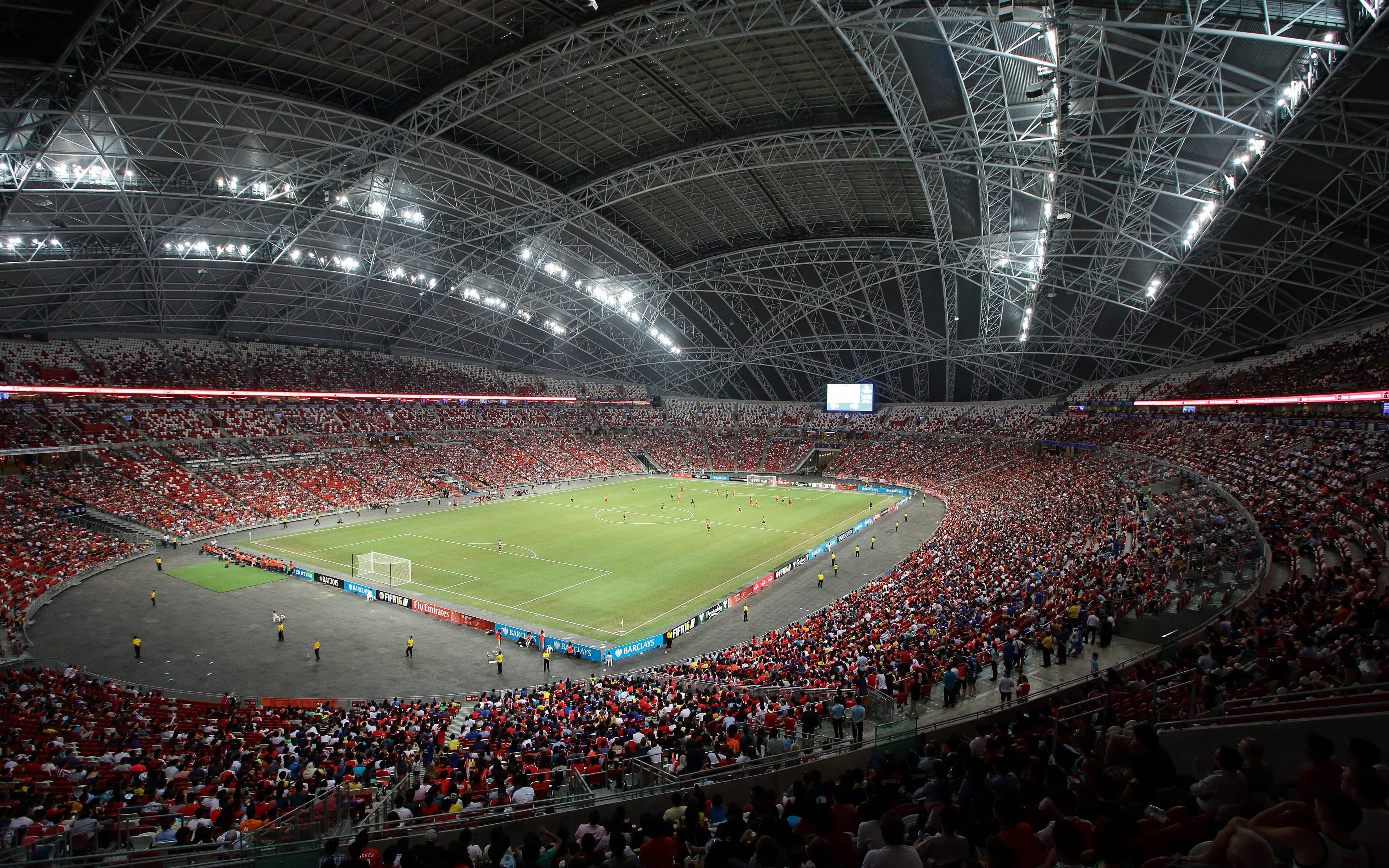 Stadium 桌布 壁紙 — 從空中俯瞰一個擠滿觀眾的足球場，巨大的鋼製屋頂下，明亮的燈光照亮綠色草坪和紅色座位。
