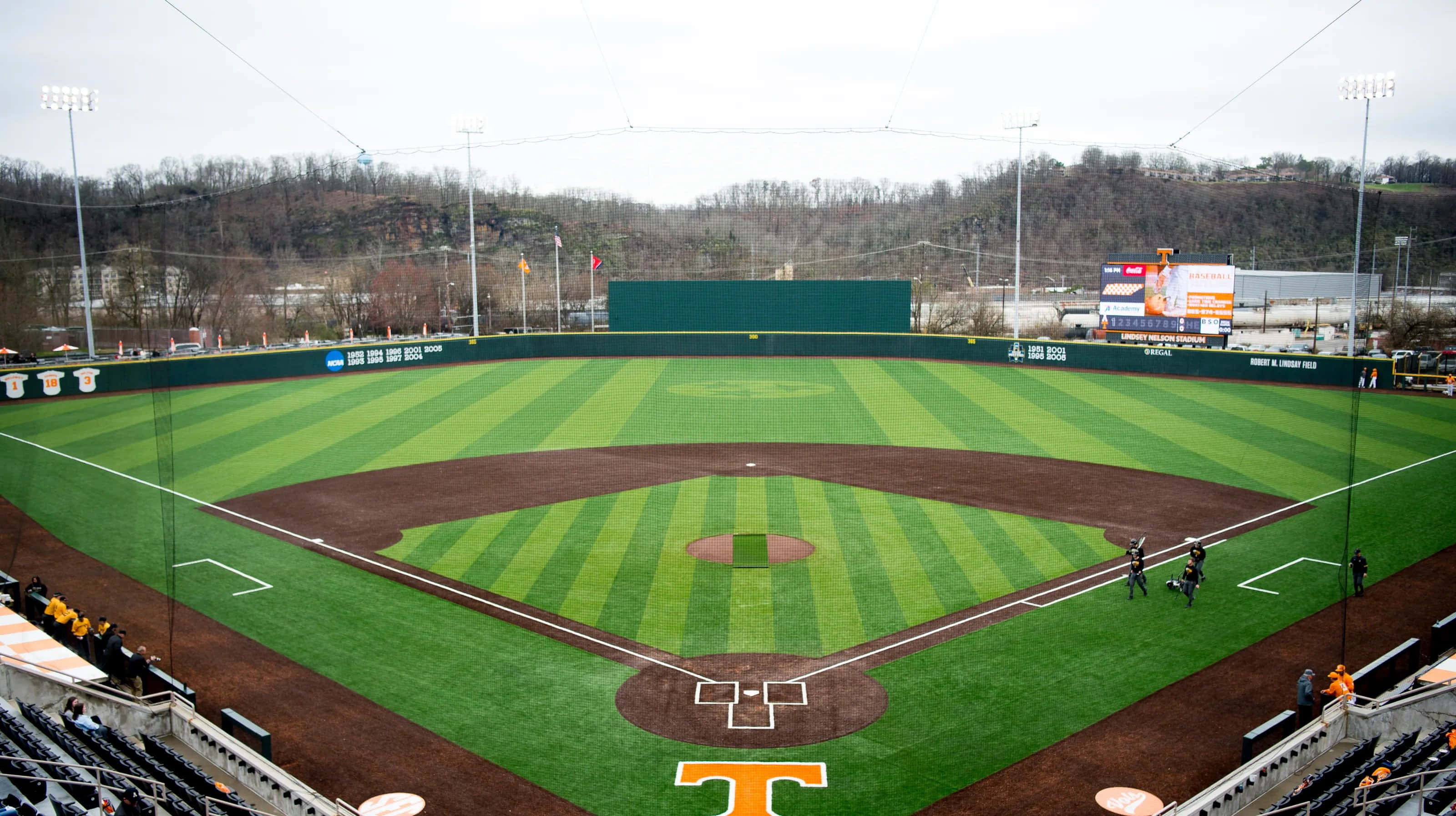 Baseball Field 桌布 壁紙 — 從空中俯瞰一個棒球場，擁有條紋狀的綠色草地和棕色泥土內野，周圍是空無一人的看台和光禿禿的樹木，天空多雲。