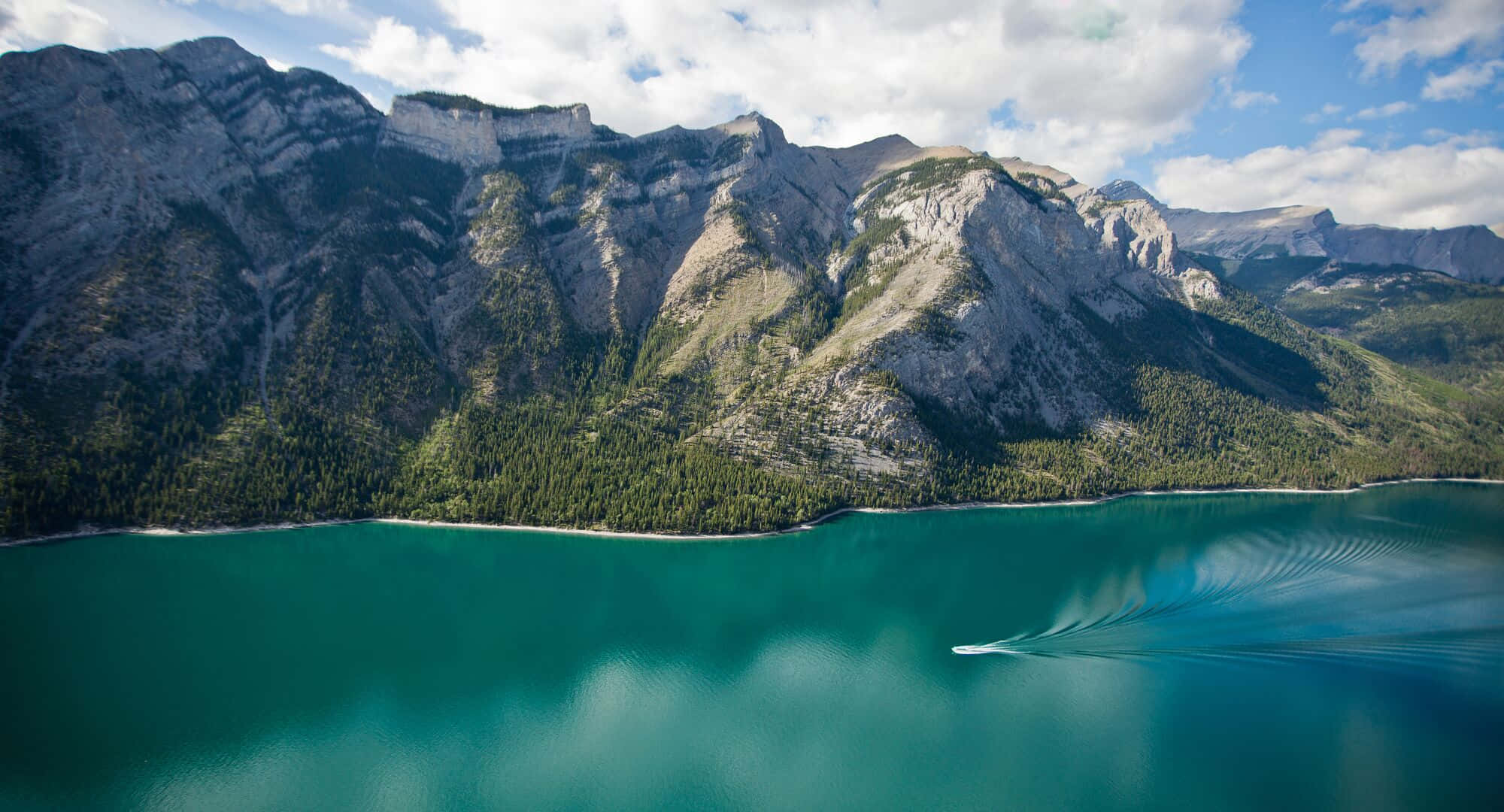 Lake 桌布 壁紙 — 從空中俯瞰一個碧綠色的湖泊，一艘快艇在部分多雲的天空下，在 towering 森林覆蓋的山脈前留下尾跡。