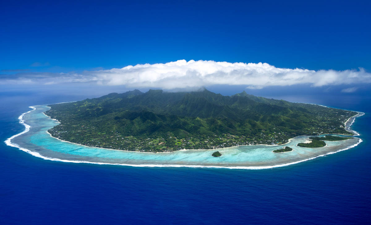 島嶼 桌布 壁紙 — 從空中俯瞰一個鬱鬱蔥蔥的綠色島嶼，擁有藍綠色潟湖和白色沙灘，被深藍色海洋環繞，天空部分多雲。