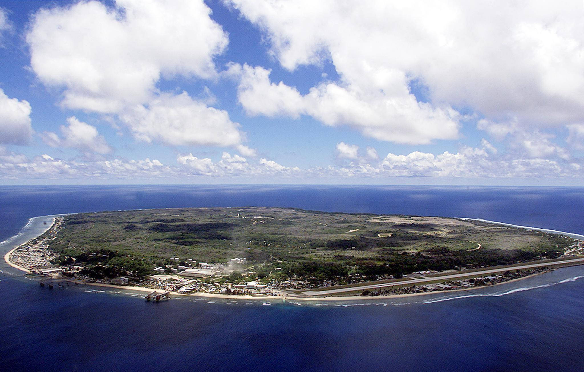 島嶼 桌布 壁紙 — 從空中俯瞰一個鬱鬱蔥蔥的綠色島嶼，島上有沿海小鎮和蜿蜒的道路，被深藍色的海洋環繞，天空部分多雲。