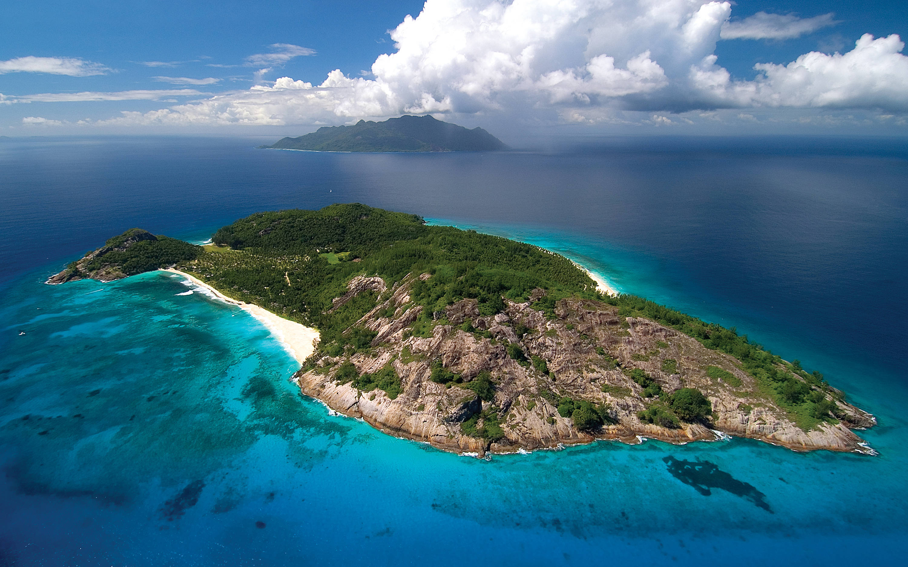 Seychelles 桌布 壁紙 — 從空中俯瞰一個鬱鬱蔥蔥的綠色島嶼，擁有白色沙灘和碧綠海水，被深藍色海洋環繞，天空部分多雲。