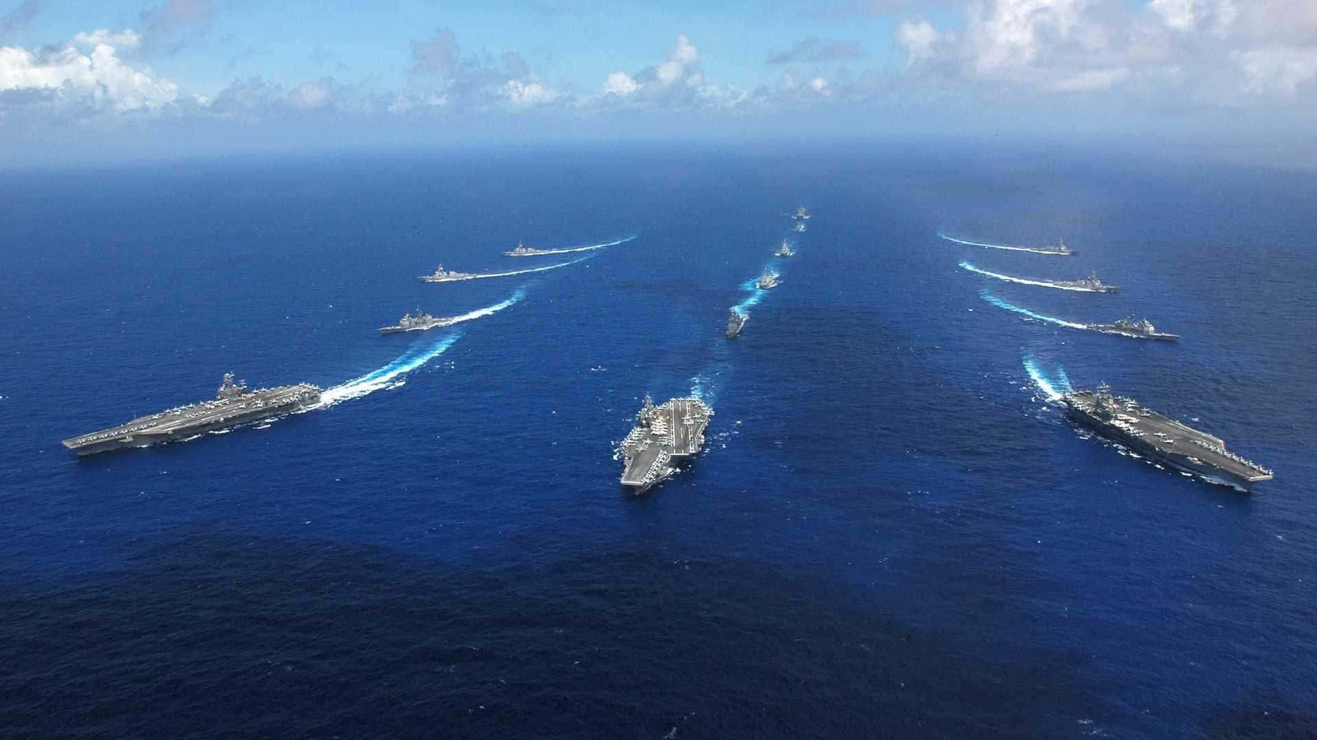 Navy 桌布 壁紙 — 從空中俯瞰一支強大的海軍艦隊，多艘航空母艦和驅逐艦在部分多雲的天空下劃過深藍色的海洋。