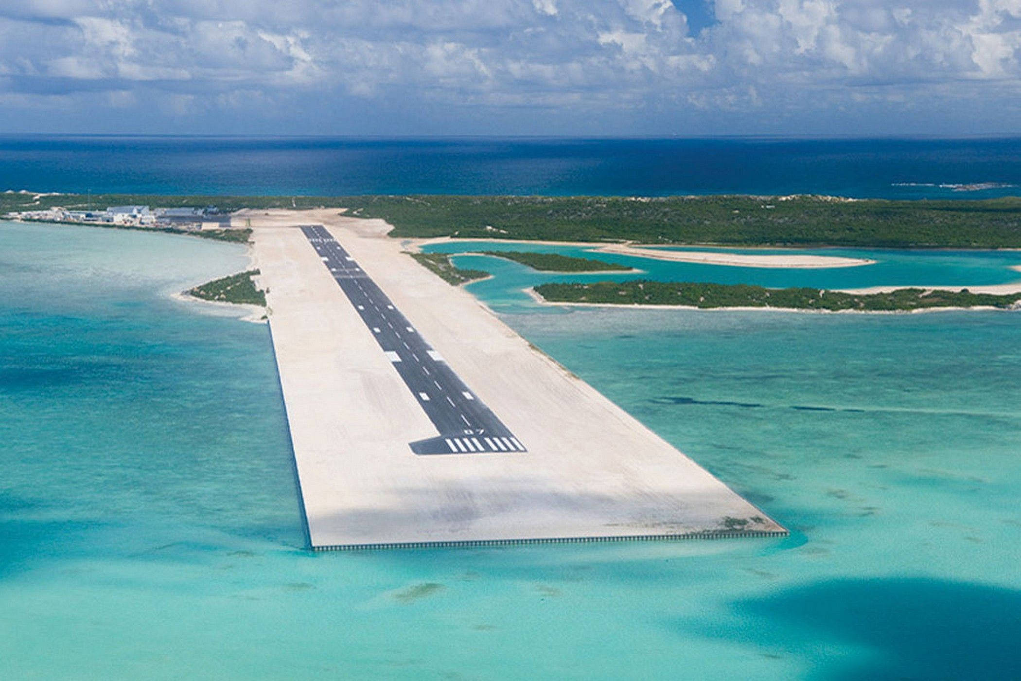 Turks And Caicos 桌布 壁紙 — 從空中俯瞰一條長長的混凝土跑道，延伸在碧綠色的海水上，通往一個鬱鬱蔥蔥的綠色島嶼，天空部分多雲。