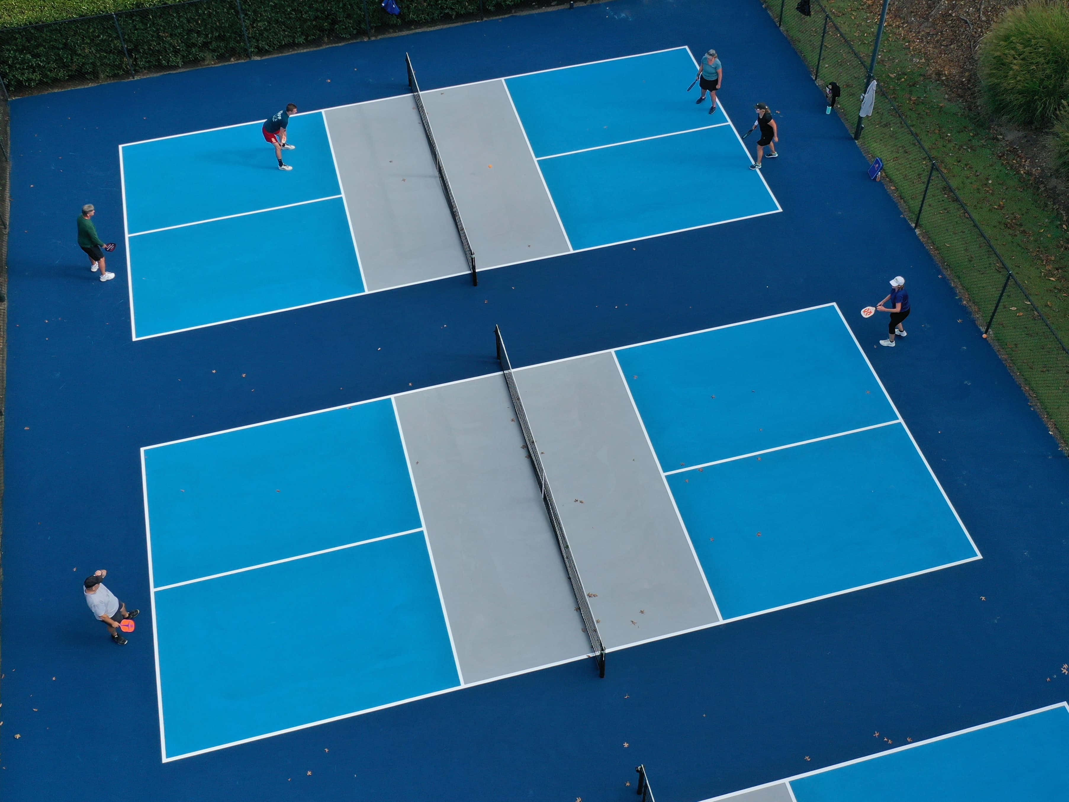 Pickleball 桌布 壁紙 — 從空中俯瞰兩個相鄰的藍色網球場，球員們在綠樹環繞中積極參與比賽。