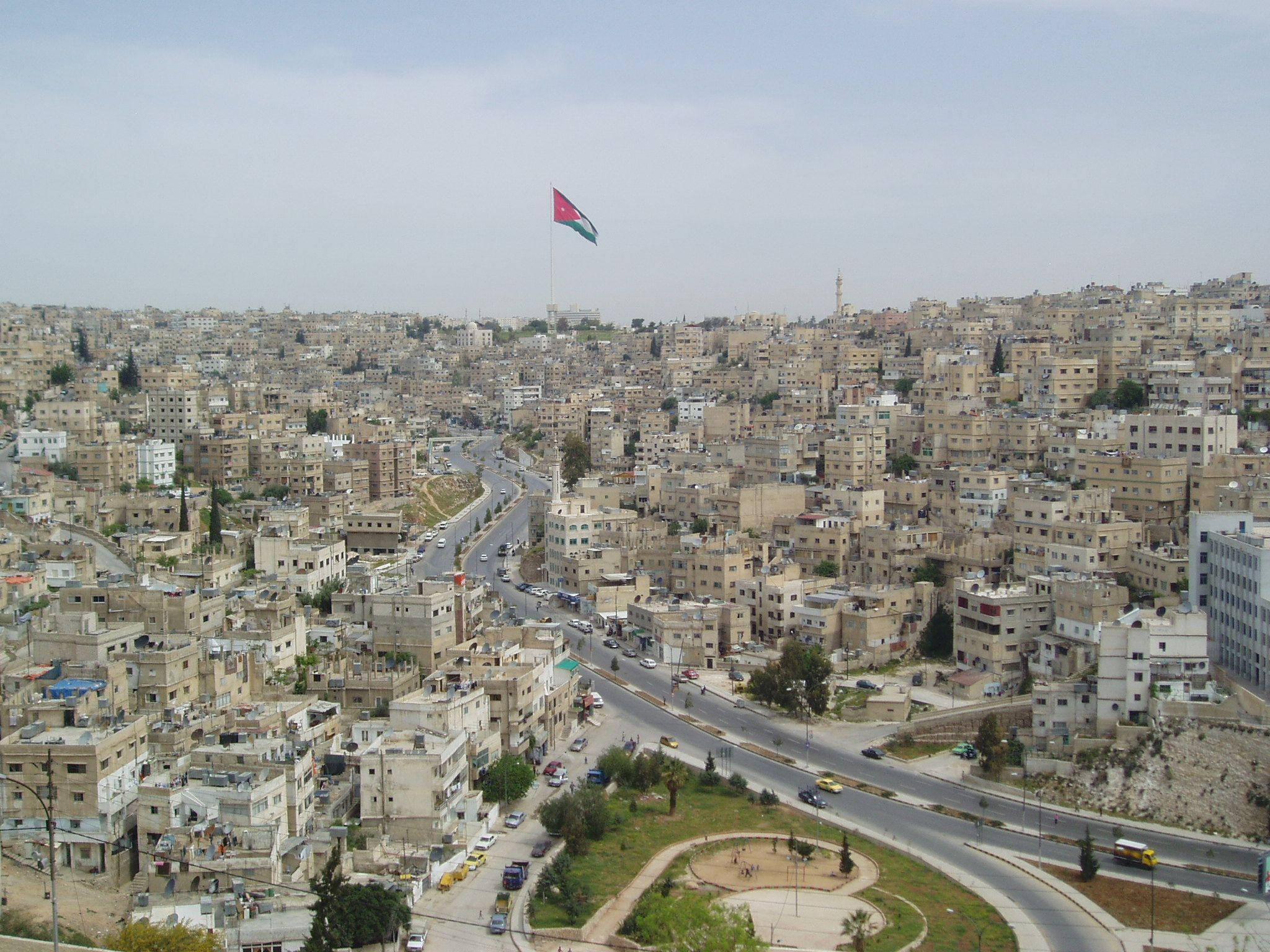 Jordan 桌布 壁紙 — 從空中俯瞰密集的城市景觀，有米色建築、蜿蜒的道路，以及在山頂上飄揚的國旗，天空朦朧。