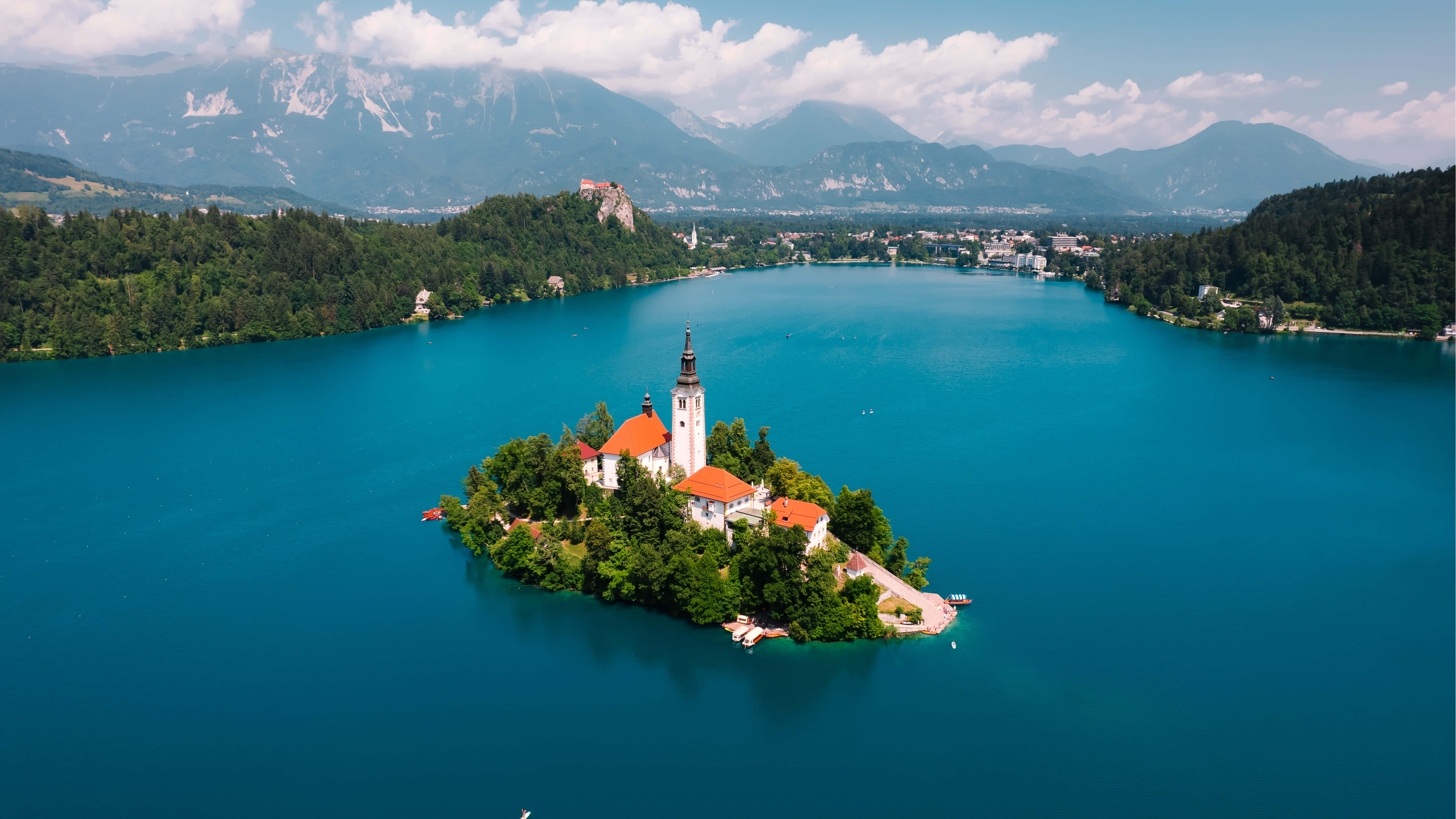 Lake Bled 桌布 壁紙 — 從空中俯瞰寧靜的碧綠湖泊，有一個小島，島上有一座白色教堂，橙色屋頂，周圍環繞著茂密的綠植和遠處積雪的山峰，天空部分多雲。