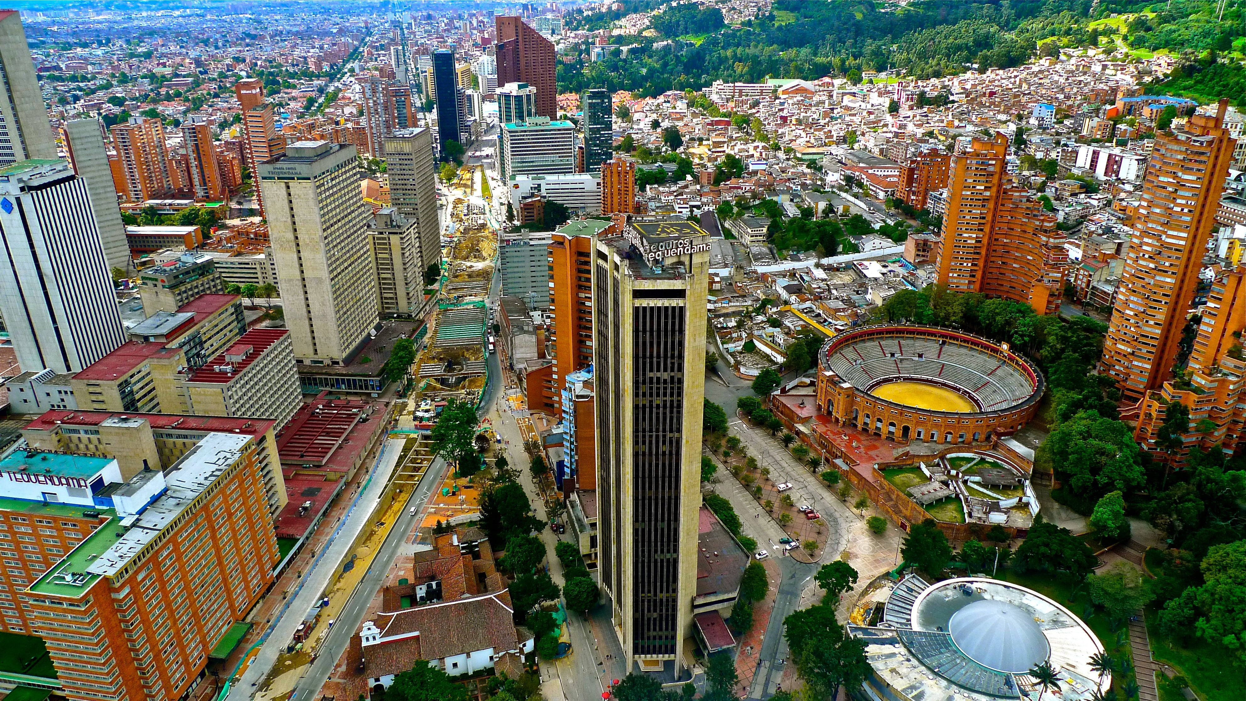 Bogota 桌布 壁紙 — 從空中俯瞰廣闊的城市景觀，包括現代摩天大樓、密集的住宅建築，以及坐落在綠色山丘中的顯著鬥牛場。
