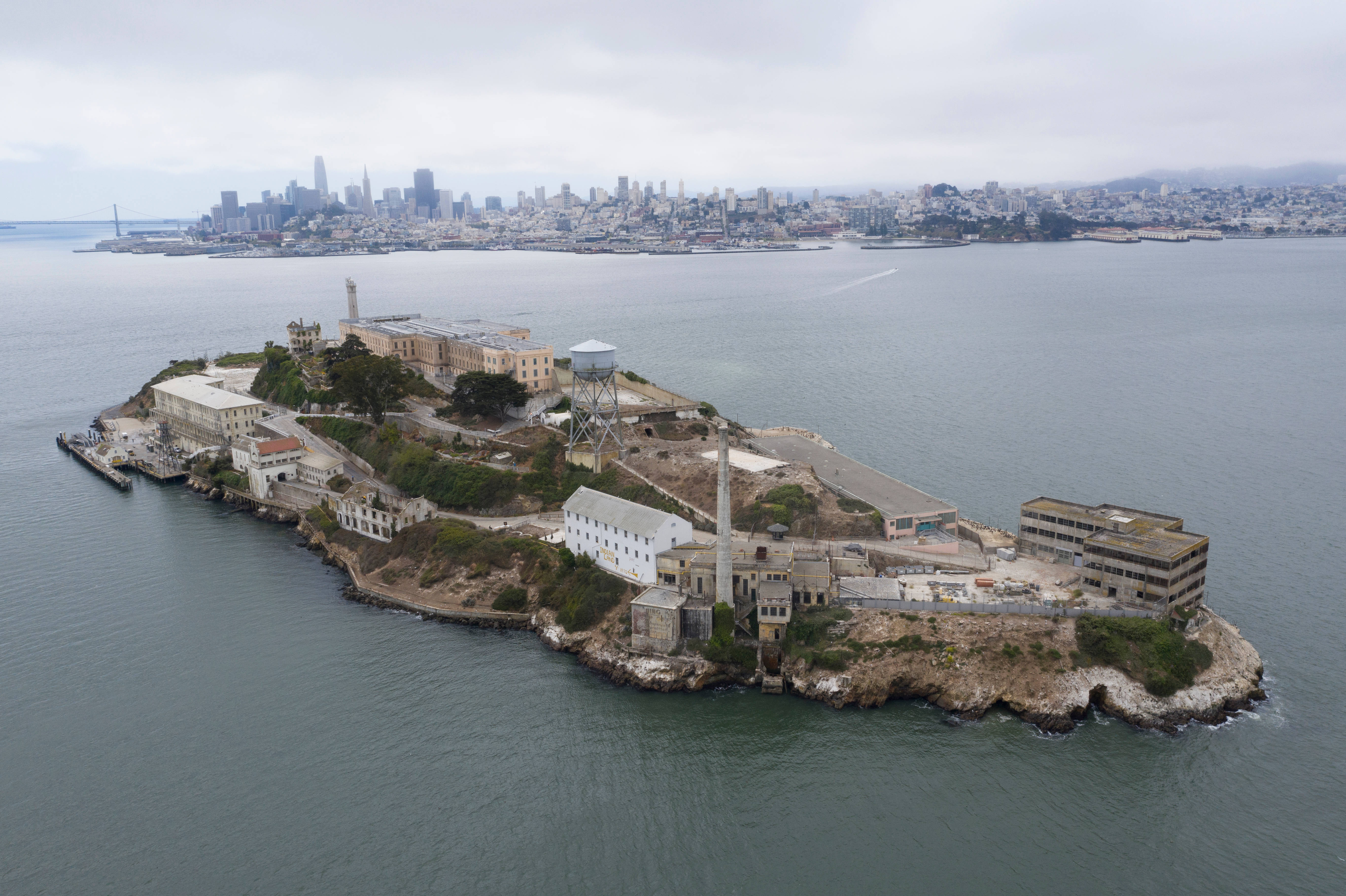 島嶼 桌布 壁紙 — 從空中俯瞰惡魔島，歷史悠久的監獄建築被灰色的海水環繞，遠處是陰天下的舊金山天際線。