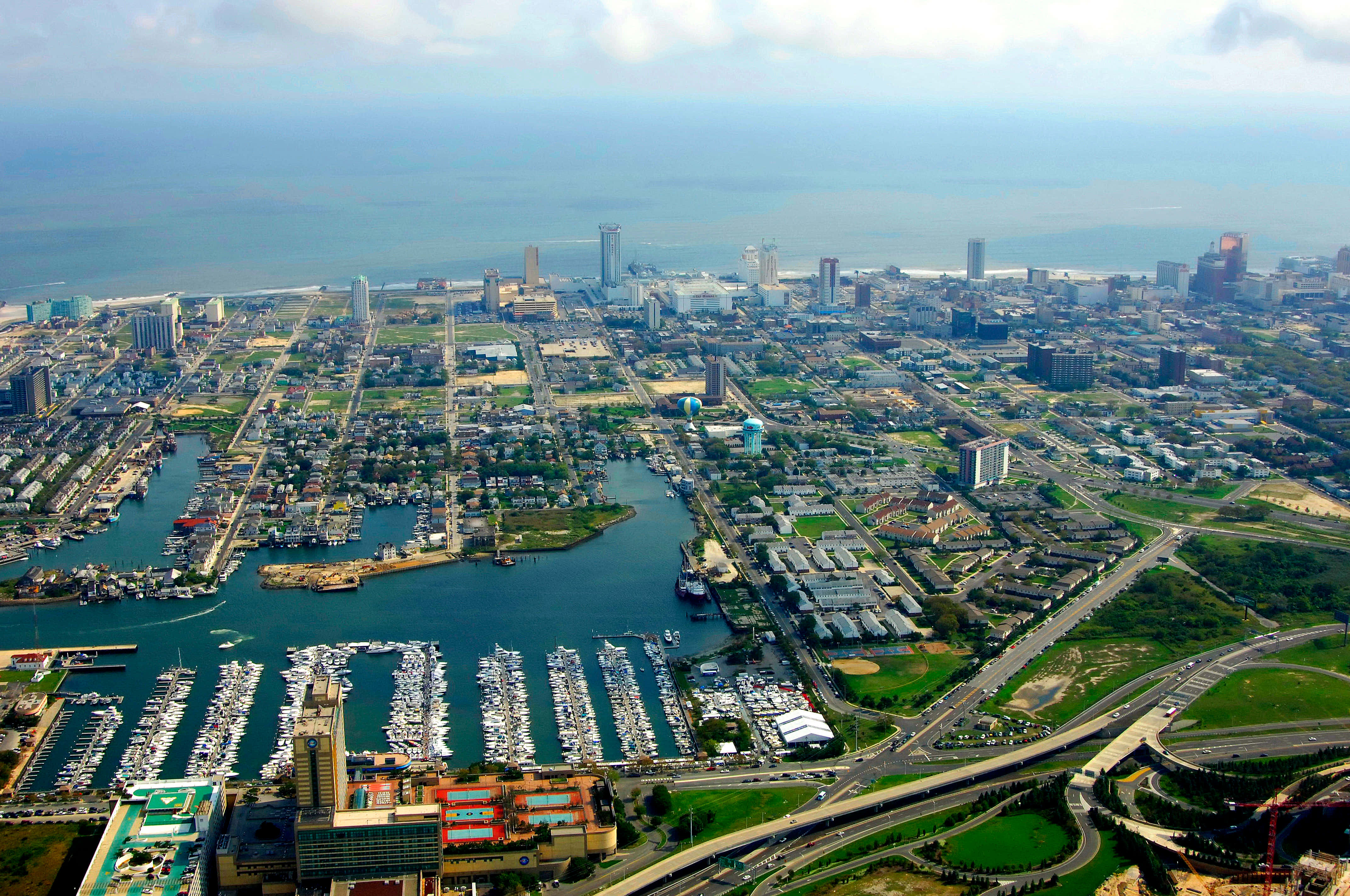 城市 桌布 壁紙 — 從空中俯瞰沿海城市，碼頭停滿船隻，現代建築在部分多雲的天空下向海洋延伸。