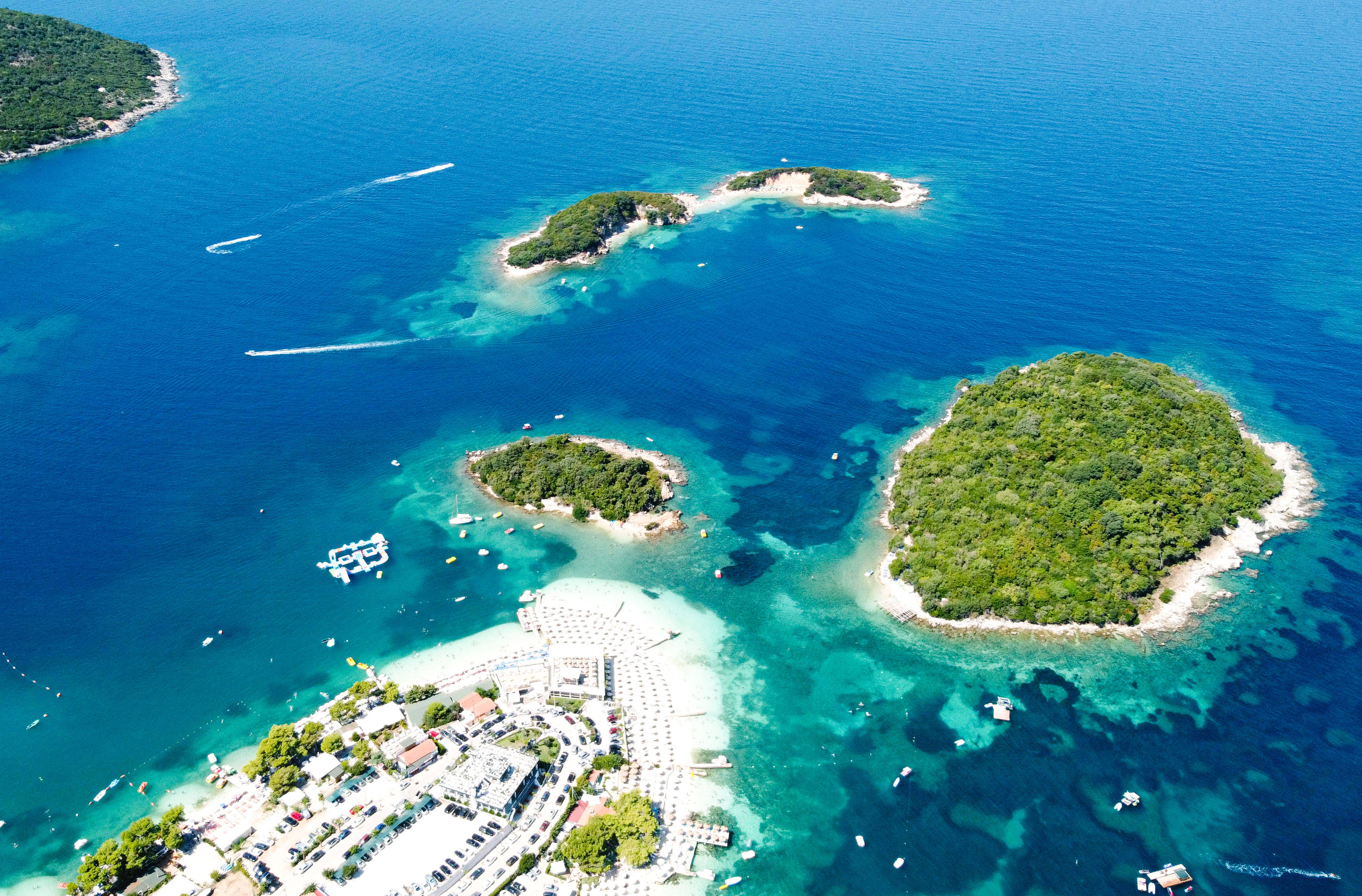 Albania 桌布 壁紙 — 從空中俯瞰碧綠海水環繞著鬱蔥的綠色島嶼，設有沿海度假村和快艇留下白色痕跡。
