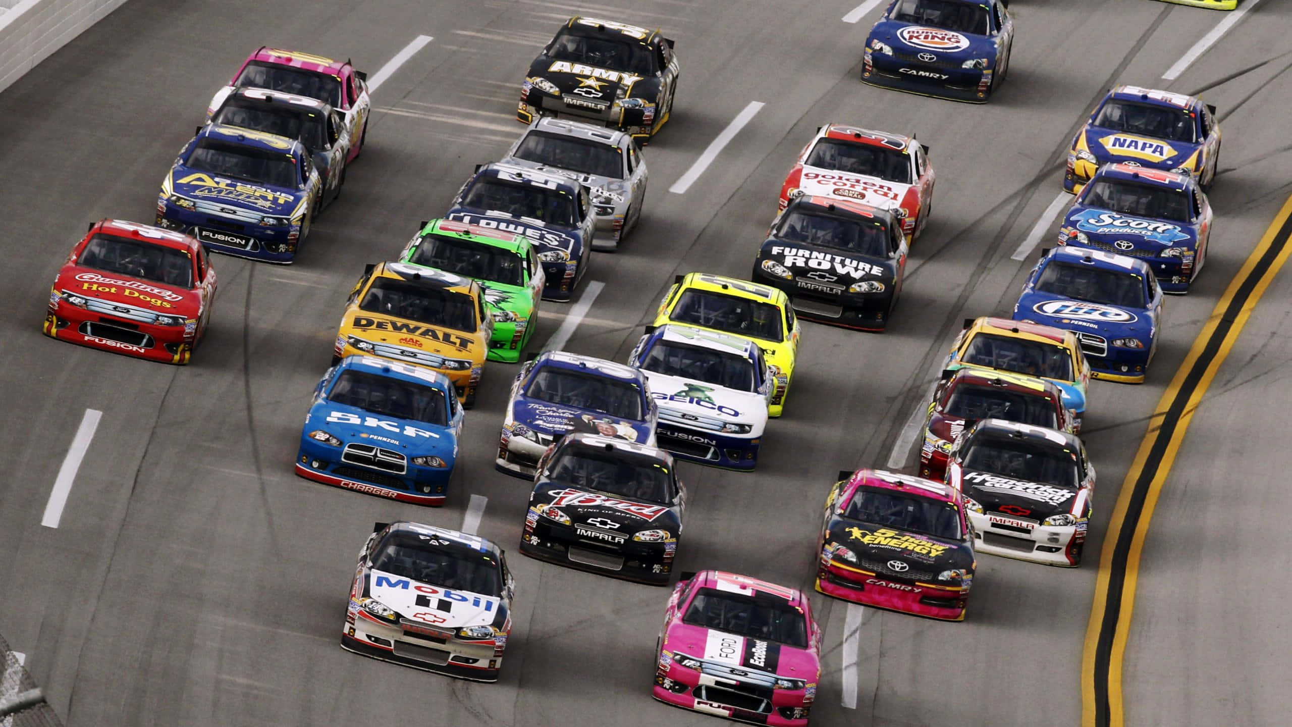 nascar 桌布 壁紙 — 從空中俯瞰色彩繽紛的NASCAR賽車緊密排列，在夜間彎曲賽道上疾馳。