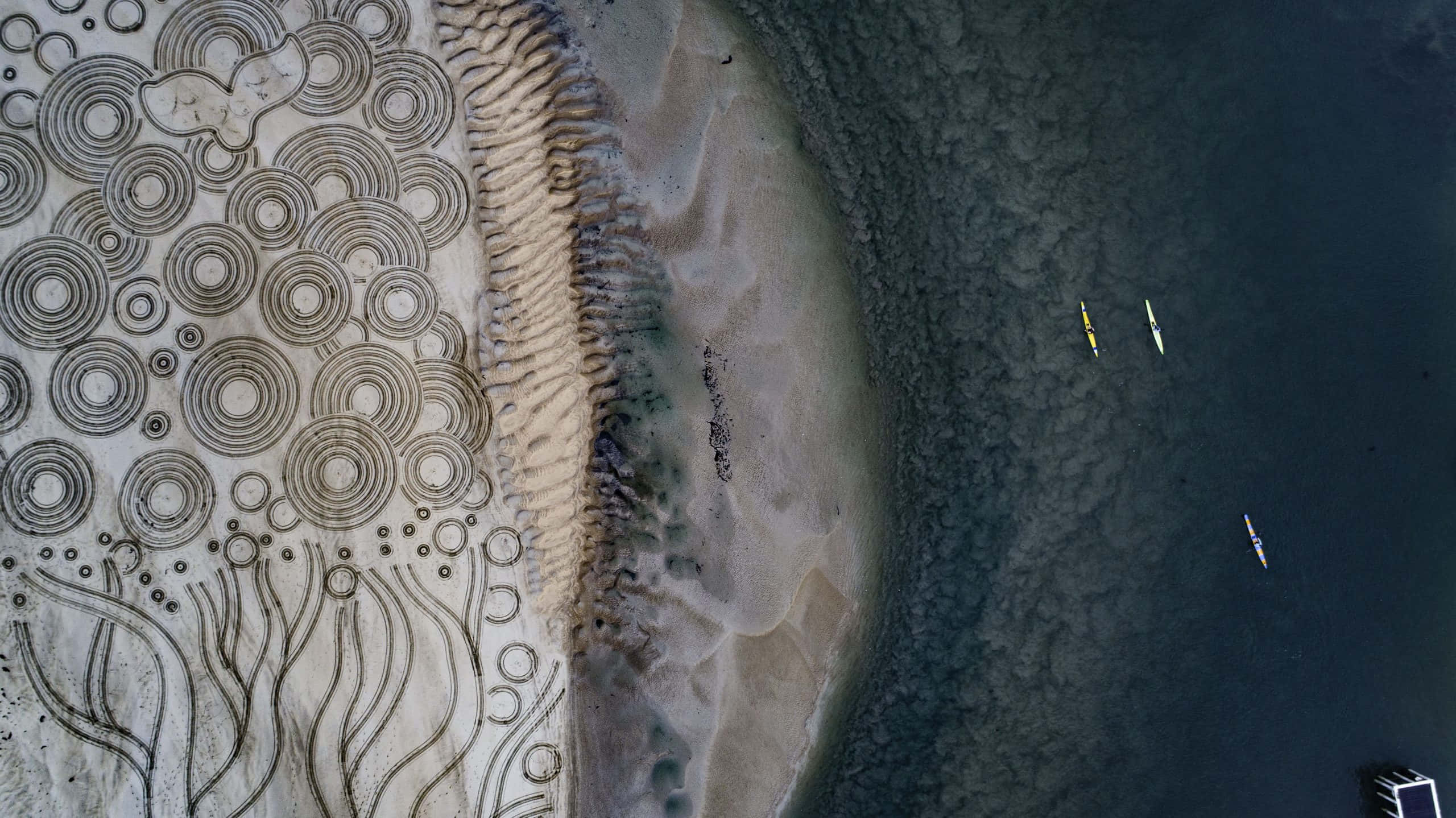 Sand Art 桌布 壁紙 — 從空中俯瞰複雜的沙藝圖案與紋理海岸線交匯，皮划艇在平靜的水面上漂浮。